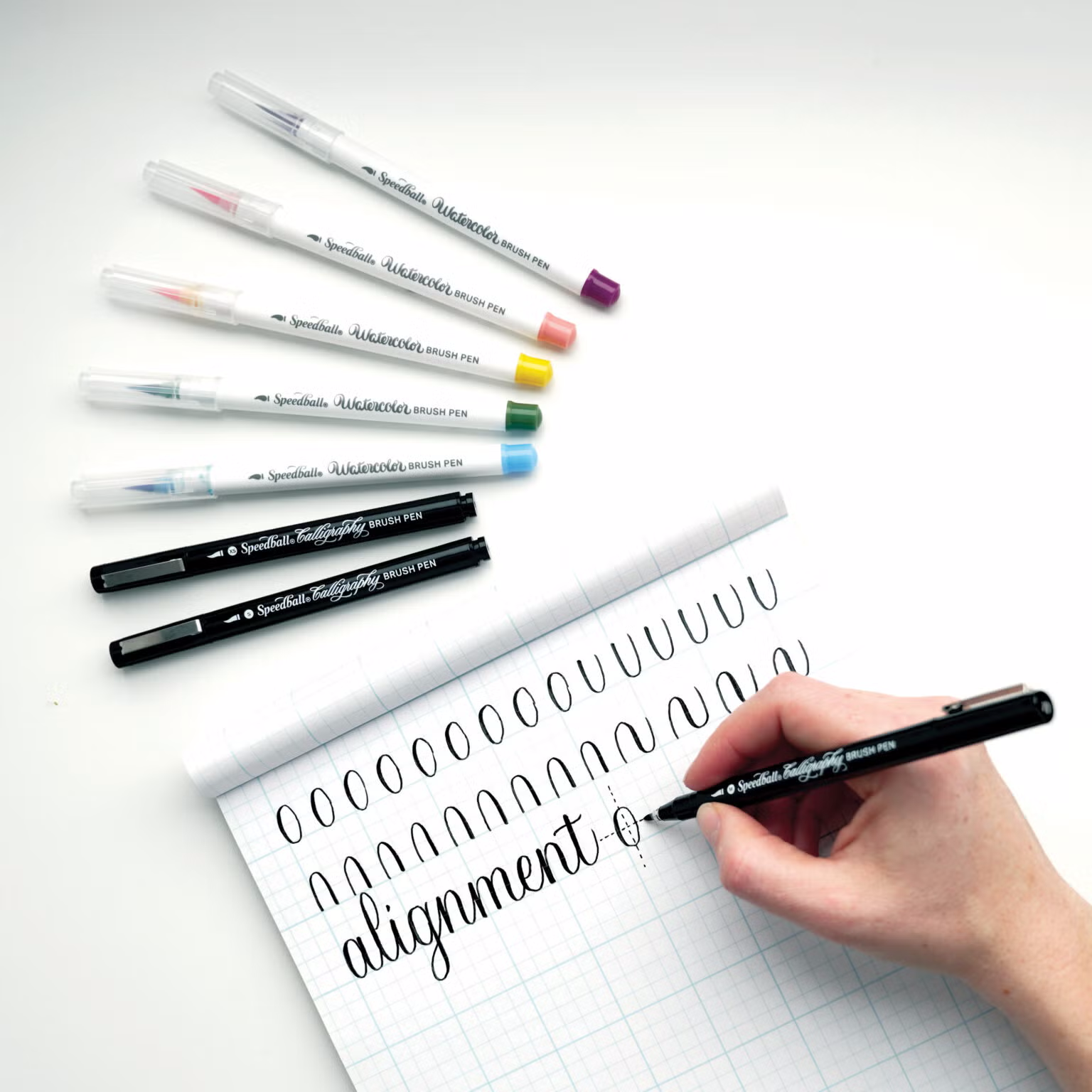 A set of Speedball hand-lettering brush markers with various colours, including black and coloured markers, laid out on a white surface with a grid paper and a pen in the process of writing.
