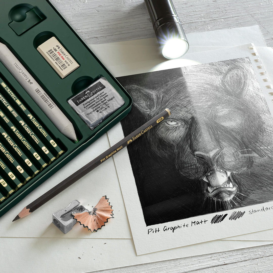 A set of Faber-Castell pencils and accessories in a tin besides a sketch of a lion on paper done half in Pitt Graphite matt pencils and half in 9000 Castell pencils. A torch shines over the drawing to show the variation in reflection across both sides.