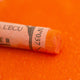 A close-up of an Orange Sennelier extra soft pastel. on the same coloured background, with the colour information printed on the paper wrap.