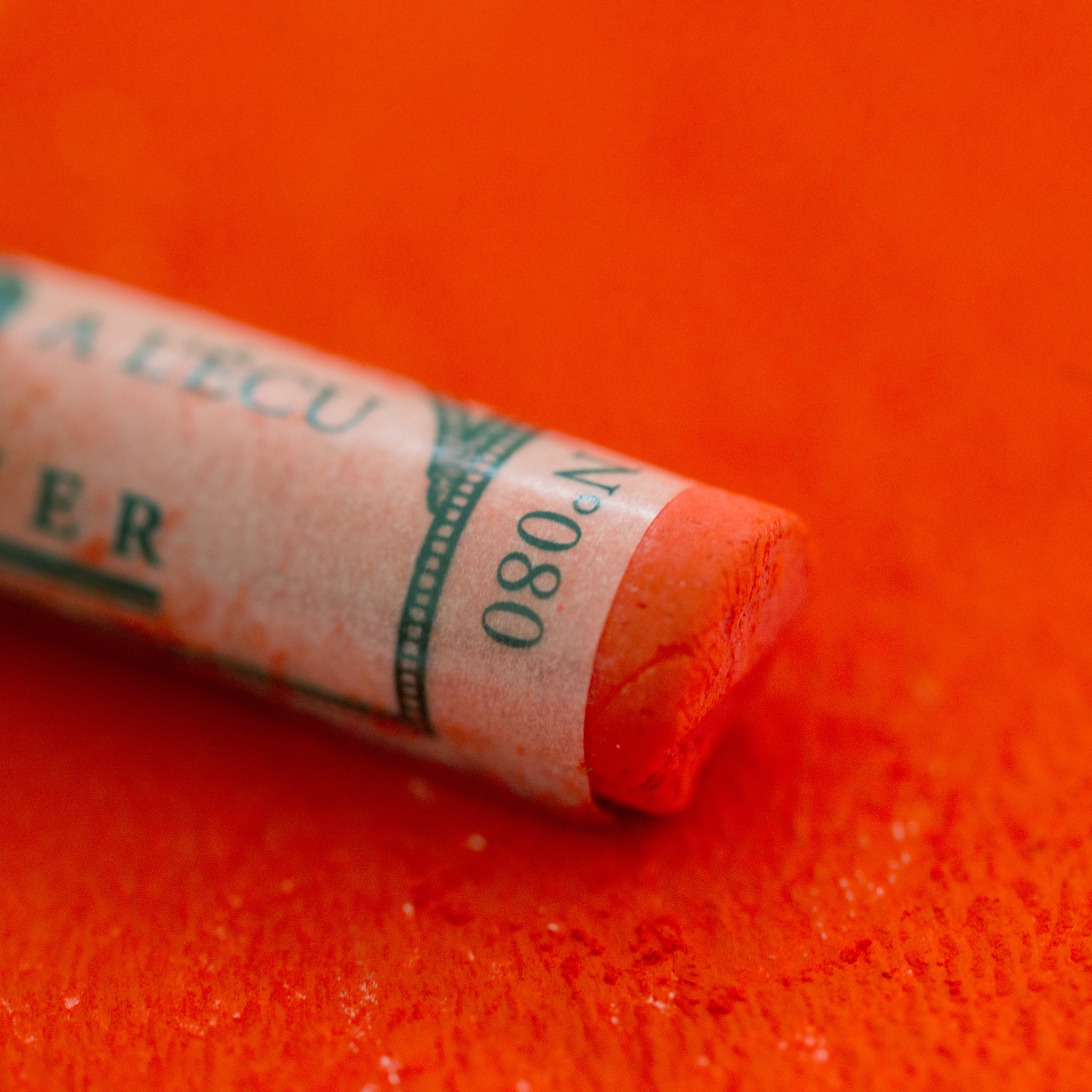 A close-up of an orange Sennelier extra soft pastel on the same coloured background, with the colour information printed on the paper wrap.