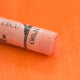 A close-up of a light orange Sennelier extra soft pastel on the same coloured background, with the colour information printed on the paper wrap.