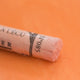 A close-up of a pale orange Sennelier extra soft pastel on the same coloured background, with the colour information printed on the paper wrap.