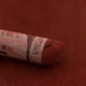 A close-up of a red-brown Sennelier extra soft pastel on the same coloured background, with the colour information printed on the paper wrap.
