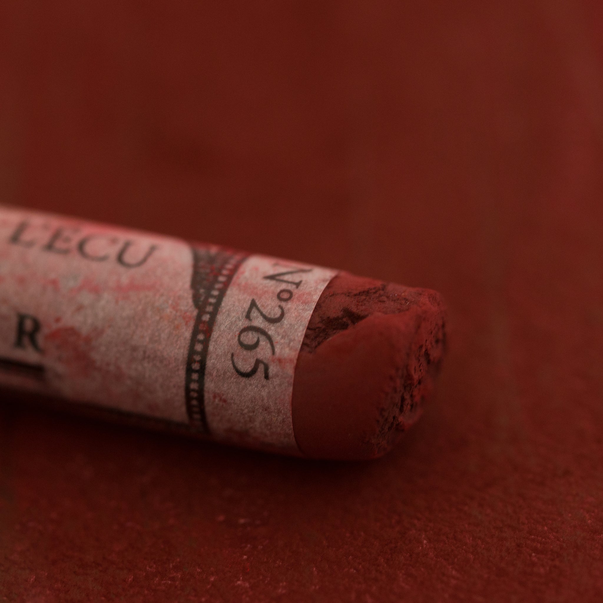 A close-up of a red-brown Sennelier extra soft pastel on the same coloured background, with the colour information printed on the paper wrap.