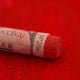 A close-up of a red Sennelier extra soft pastel on the same coloured background, with the colour information printed on the paper wrap.