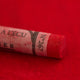 A close-up of a red Sennelier extra soft pastel on the same coloured background, with the colour information printed on the paper wrap.