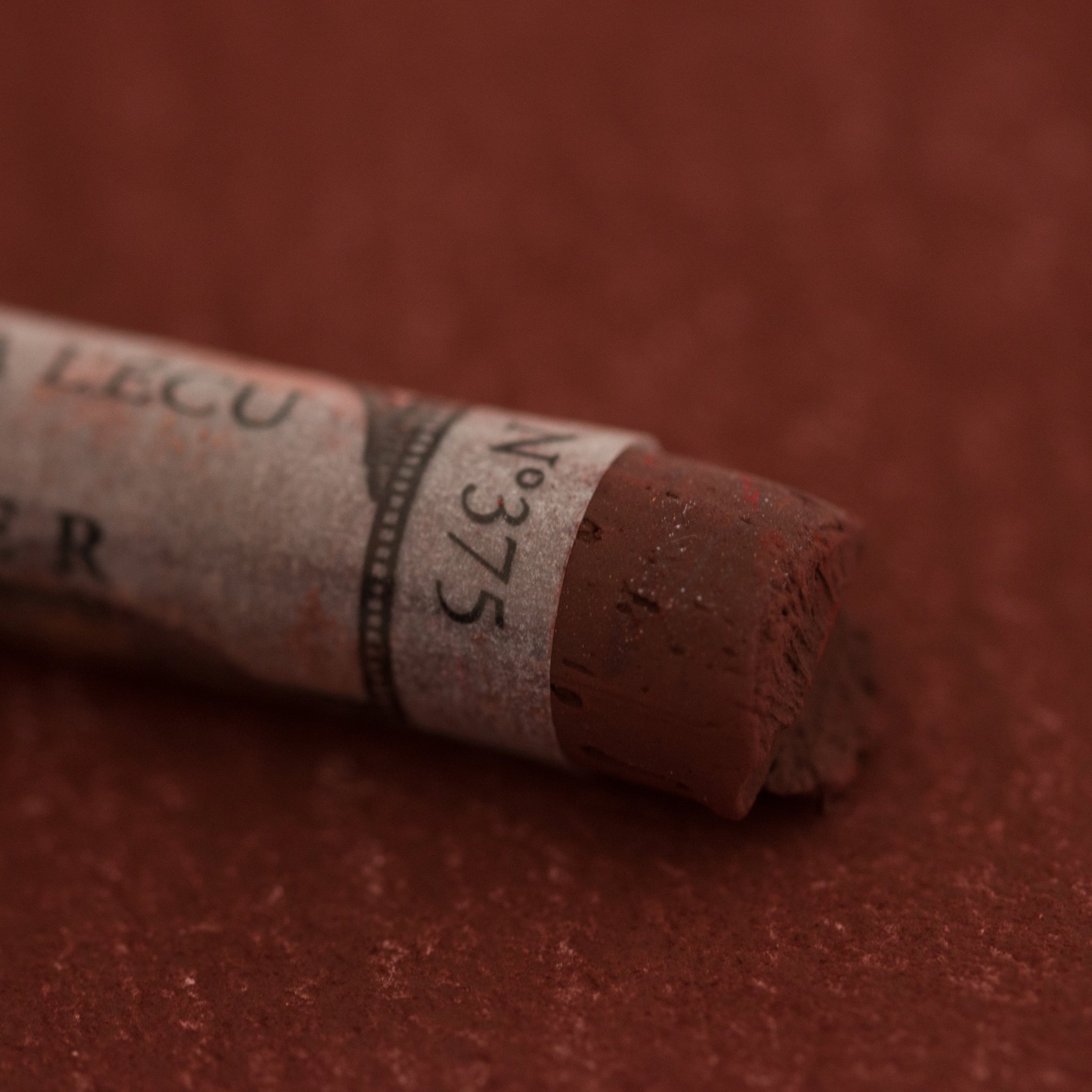 A close-up of a red-brown Sennelier extra soft pastel on the same coloured background, with the colour information printed on the paper wrap.