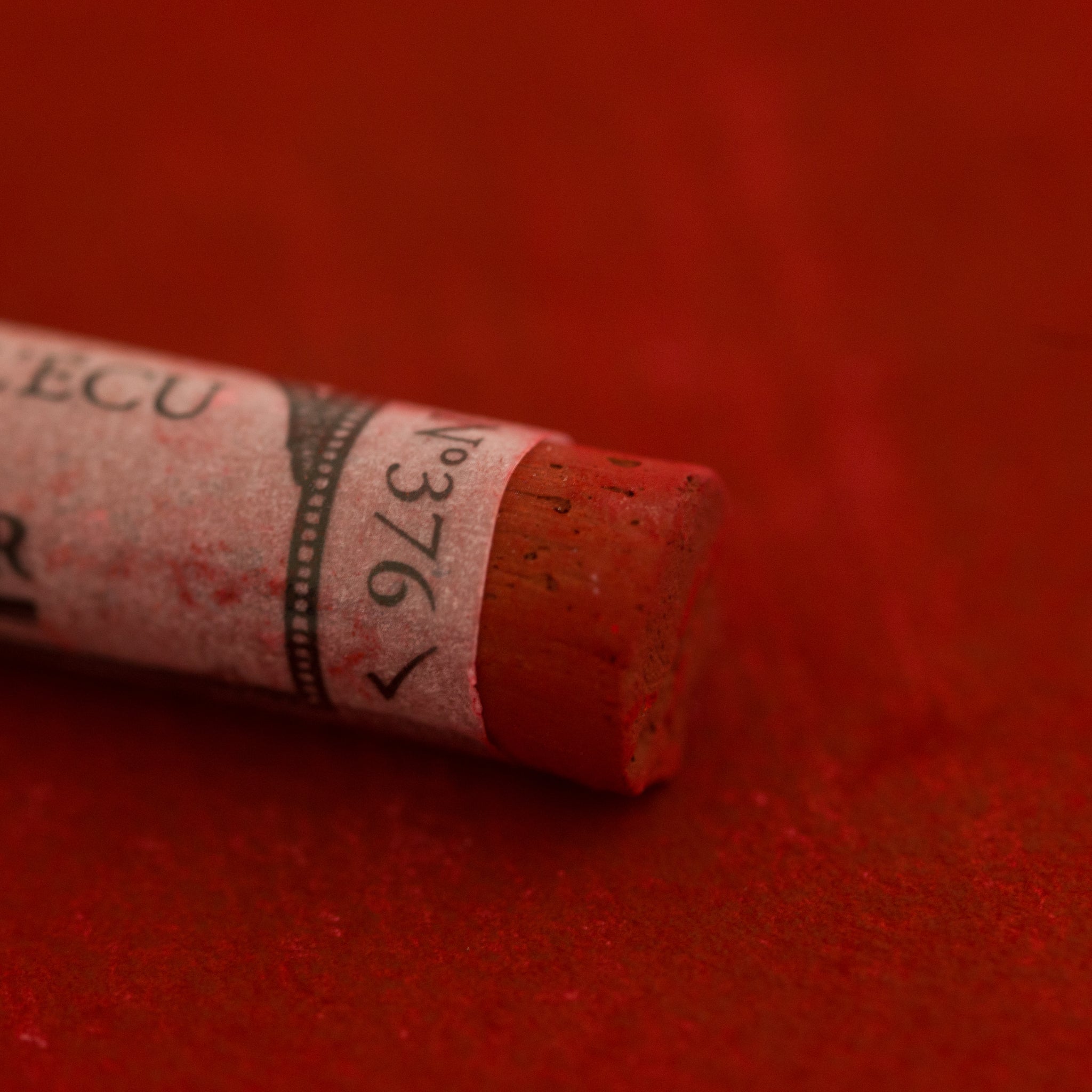 A close-up of a red-brown Sennelier extra soft pastel on the same coloured background, with the colour information printed on the paper wrap