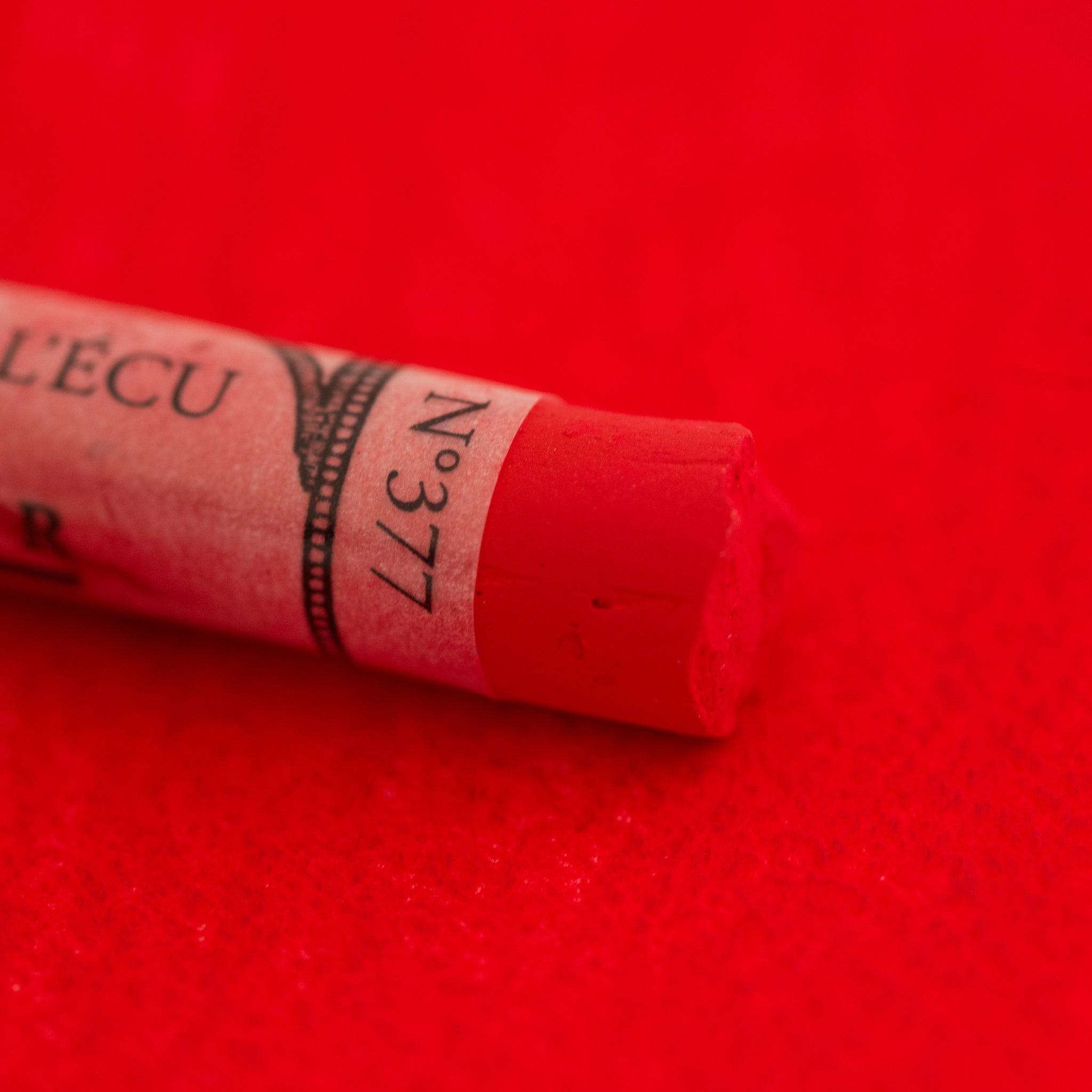 A close-up of a bright red Sennelier extra soft pastel on the same coloured background, with the colour information printed on the paper wrap.