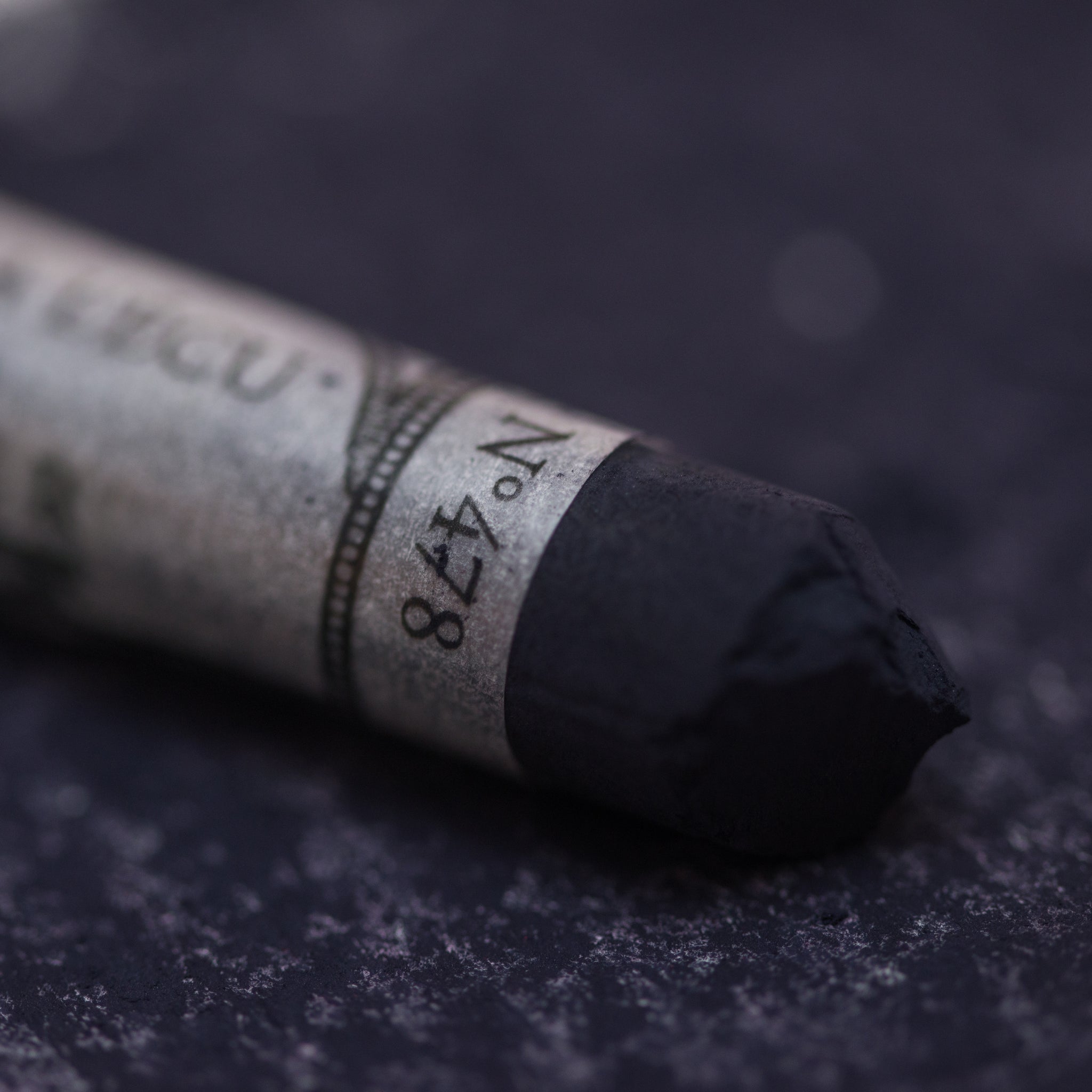 A close-up of a dark grey Sennelier extra soft pastel. on the same coloured background, with the colour information printed on the paper wrap.