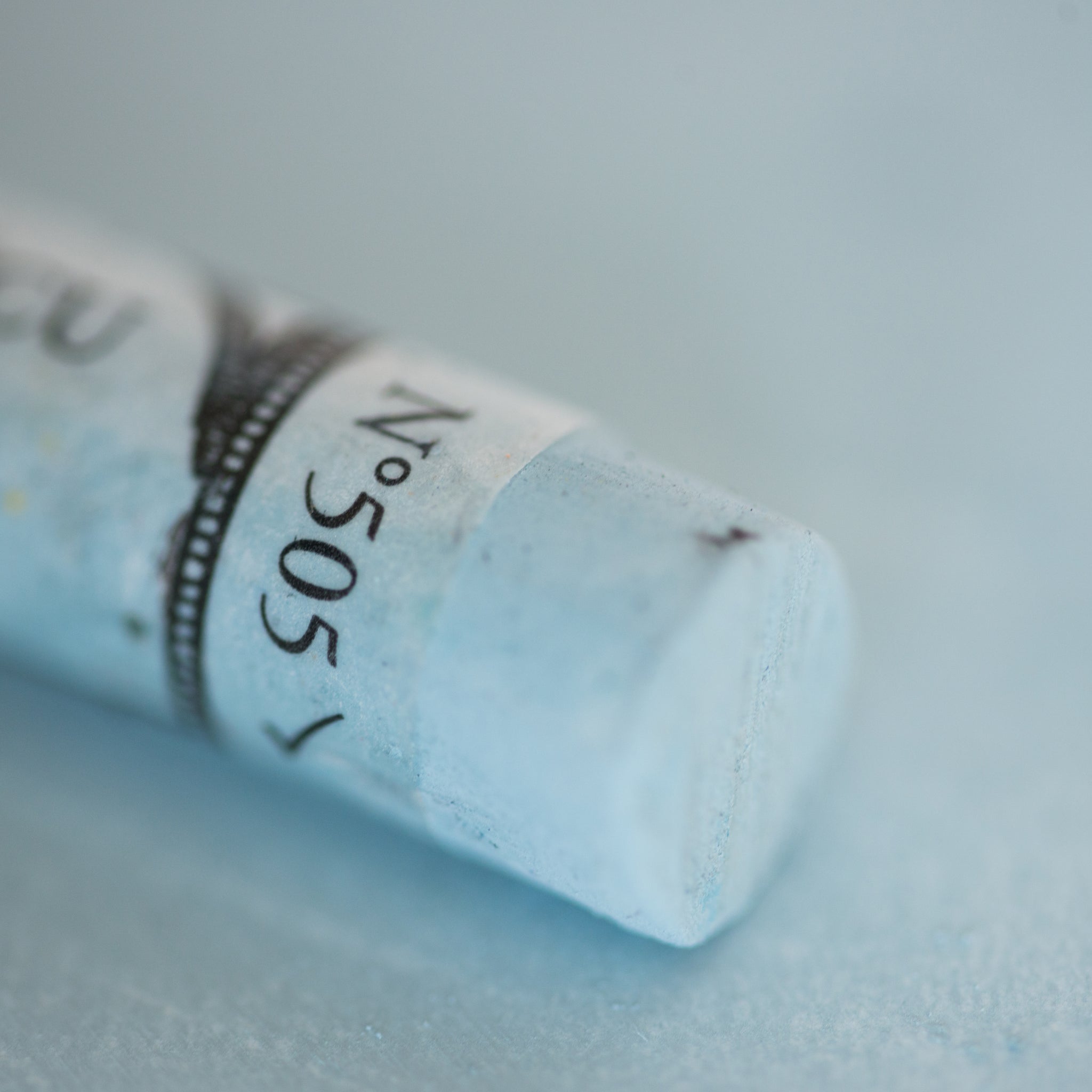 A close-up of a light blue Sennelier extra soft pastel. on the same coloured background, with the colour information printed on the paper wrap.