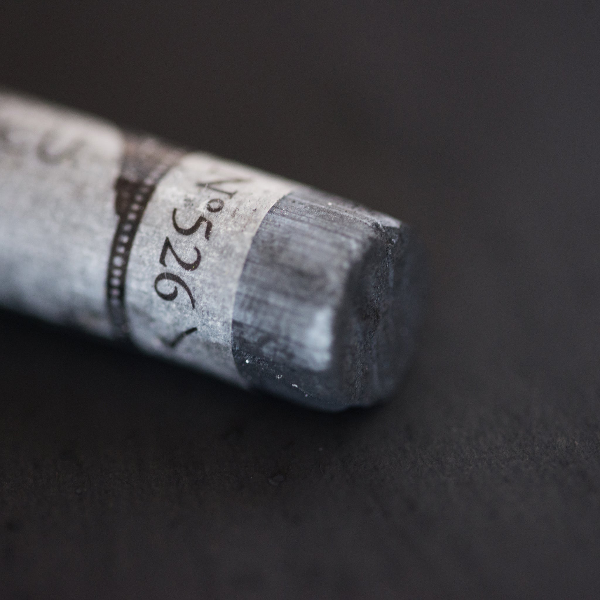 A close-up of a black Sennelier extra soft pastel. on the same coloured background, with the colour information printed on the paper wrap.
