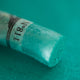 A close-up of an iridescent green Sennelier extra soft pastel. on the same coloured background, with the colour information printed on the paper wrap.