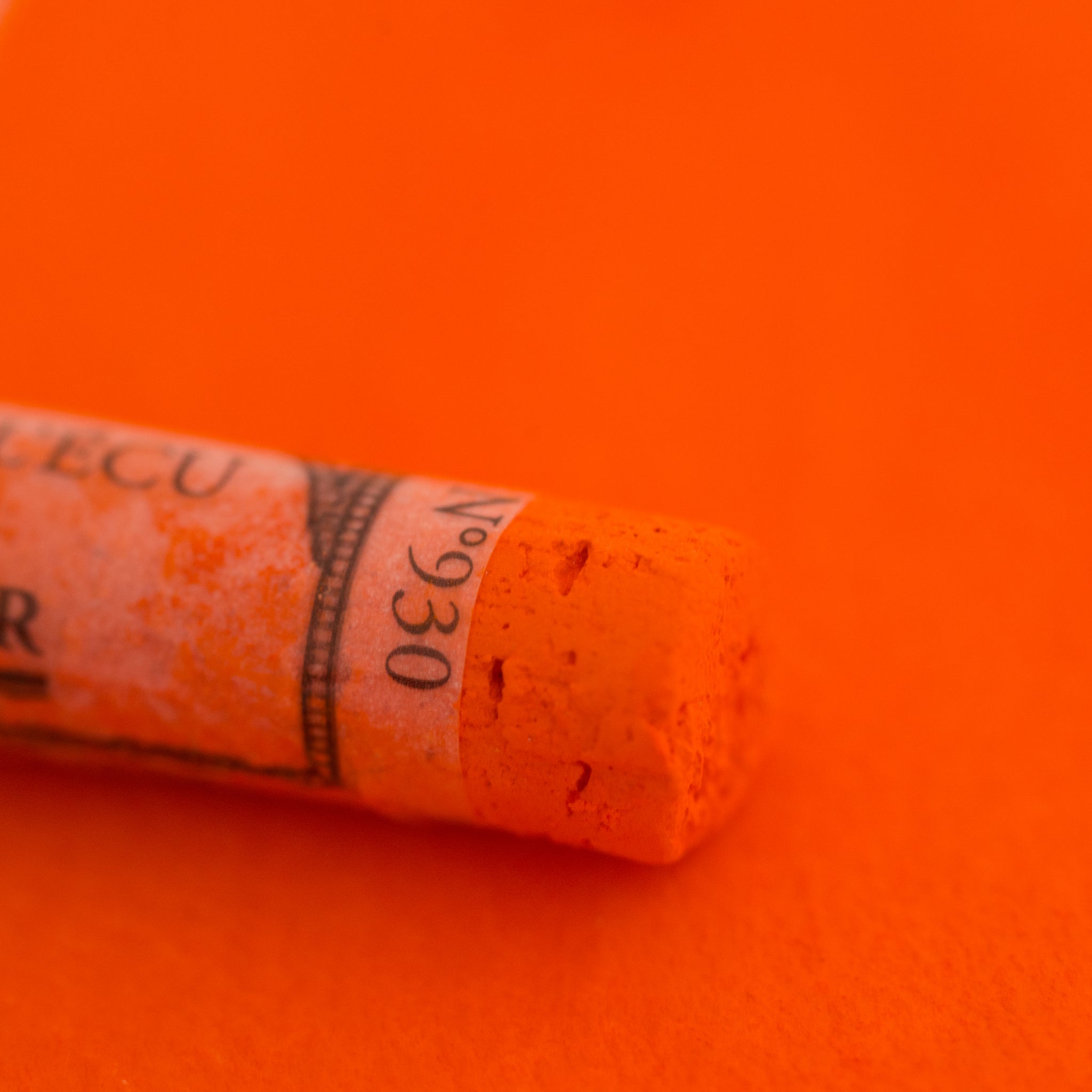 A close-up of an orange Sennelier extra soft pastel on the same coloured background, with the colour information printed on the paper wrap.