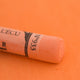 A close-up of a light orange Sennelier extra soft pastel on the same coloured background, with the colour information printed on the paper wrap.