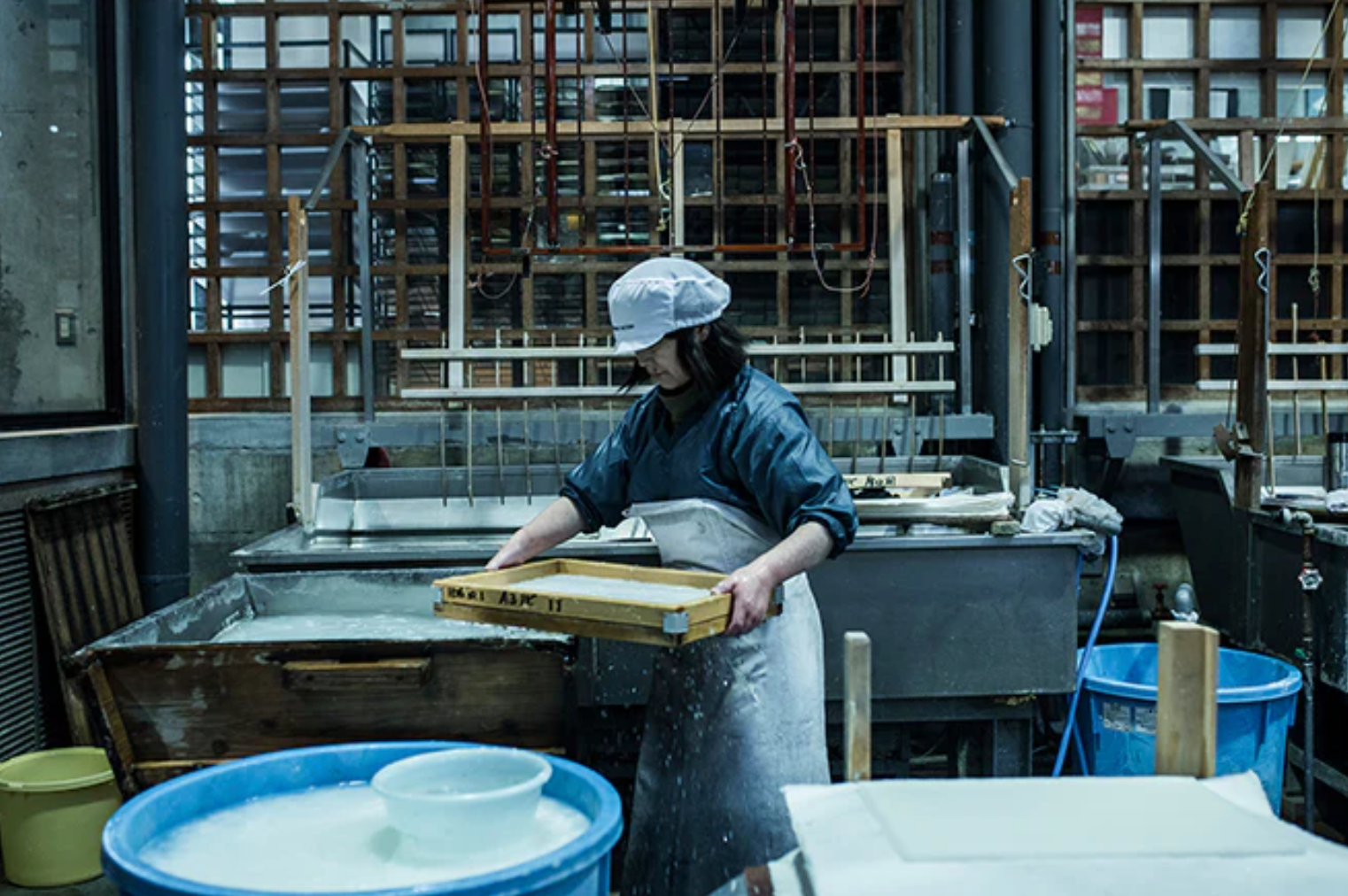 worker in a paper factory holding a frame containing paper pulp surrounded by a range of papermaking equipment