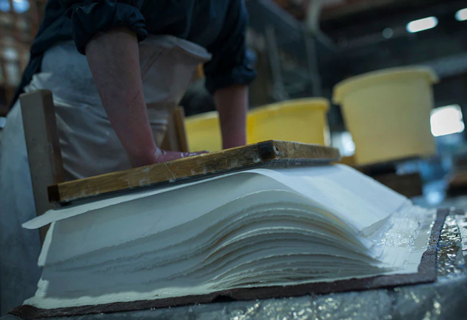 A stack of wet, freshly made paper being squashed down with a wooden tool with water spilling out of the paper stack.