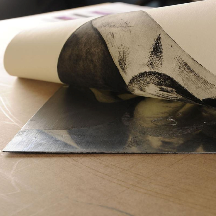 A piece of Canson Editioning paper being pull back of a metallic etched plate revealing a black print on the paper
