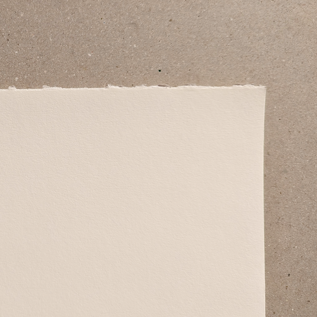 A smooth, warm-white rag paper corner with one deckled edge, placed on a neutral surface.