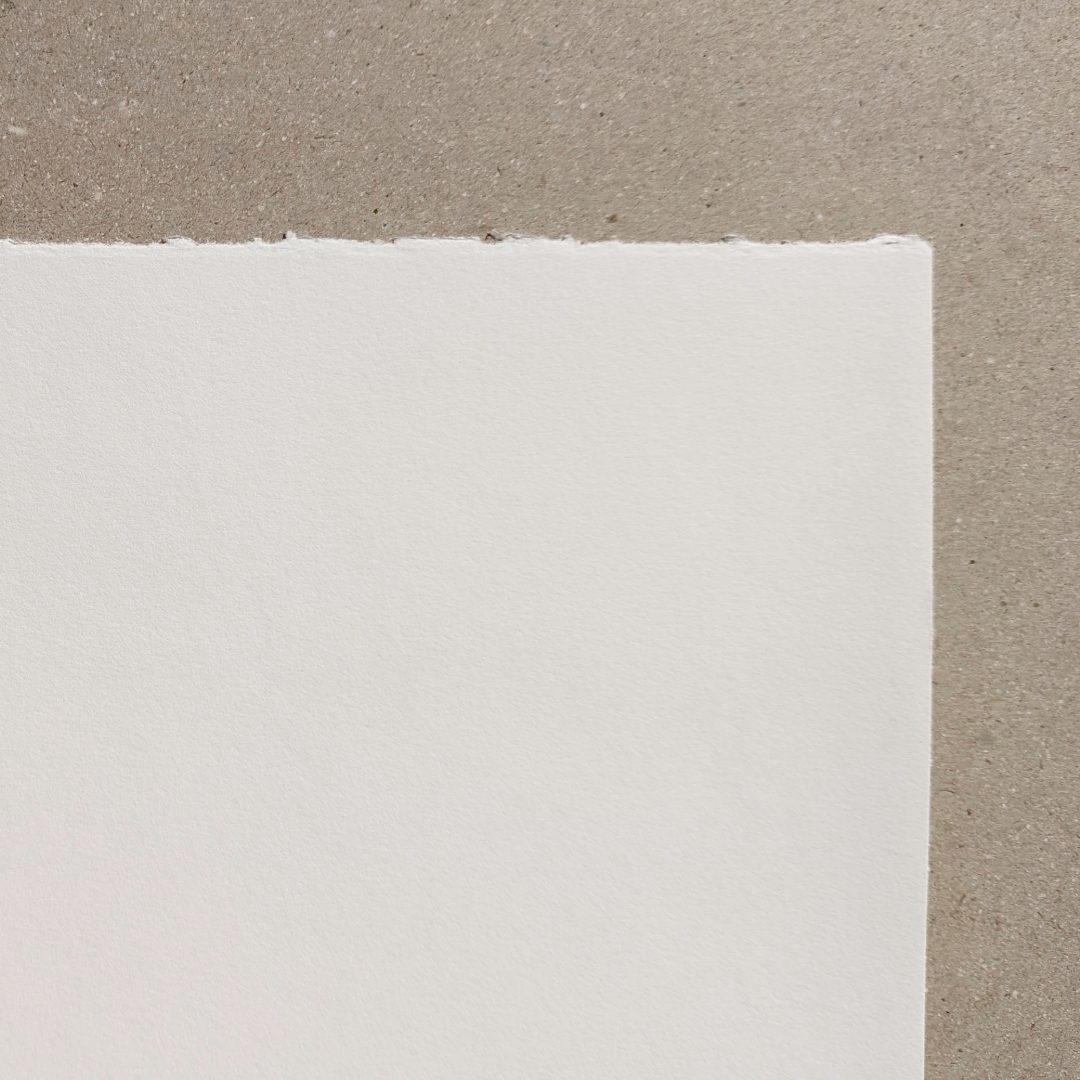 A close-up of a white paper corner with a deckle edge on one side and a light texture visible, placed against a neutral background.