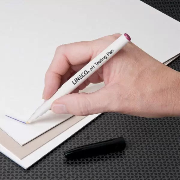 A hand holding a Lineco pH Testing Pen, drawing a line on a piece of paper, with a black mark at the end of the pen.