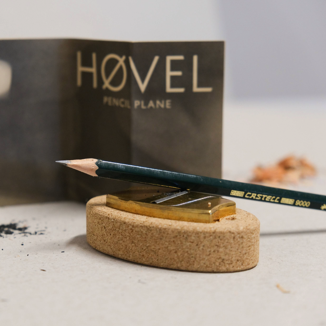 A Hovel Pencil Plane set in a cork base with a pencil resting on top of it and shavings pictured in the background.