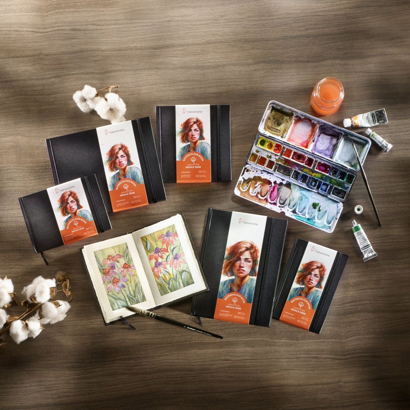 Art supplies including watercolour sets and Hahnemühle deckled edge watercolour books on a wooden surface with cotton branches.