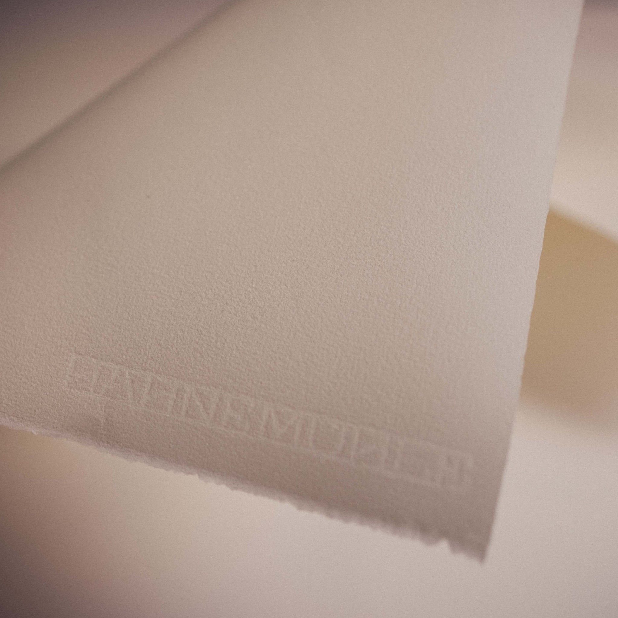 A close-up image of a piece of warm white Hahnemühle etching paper showing its texture and deckled edges. A watermark saying 'Hahnemuhle' is along the corner edge of the paper.