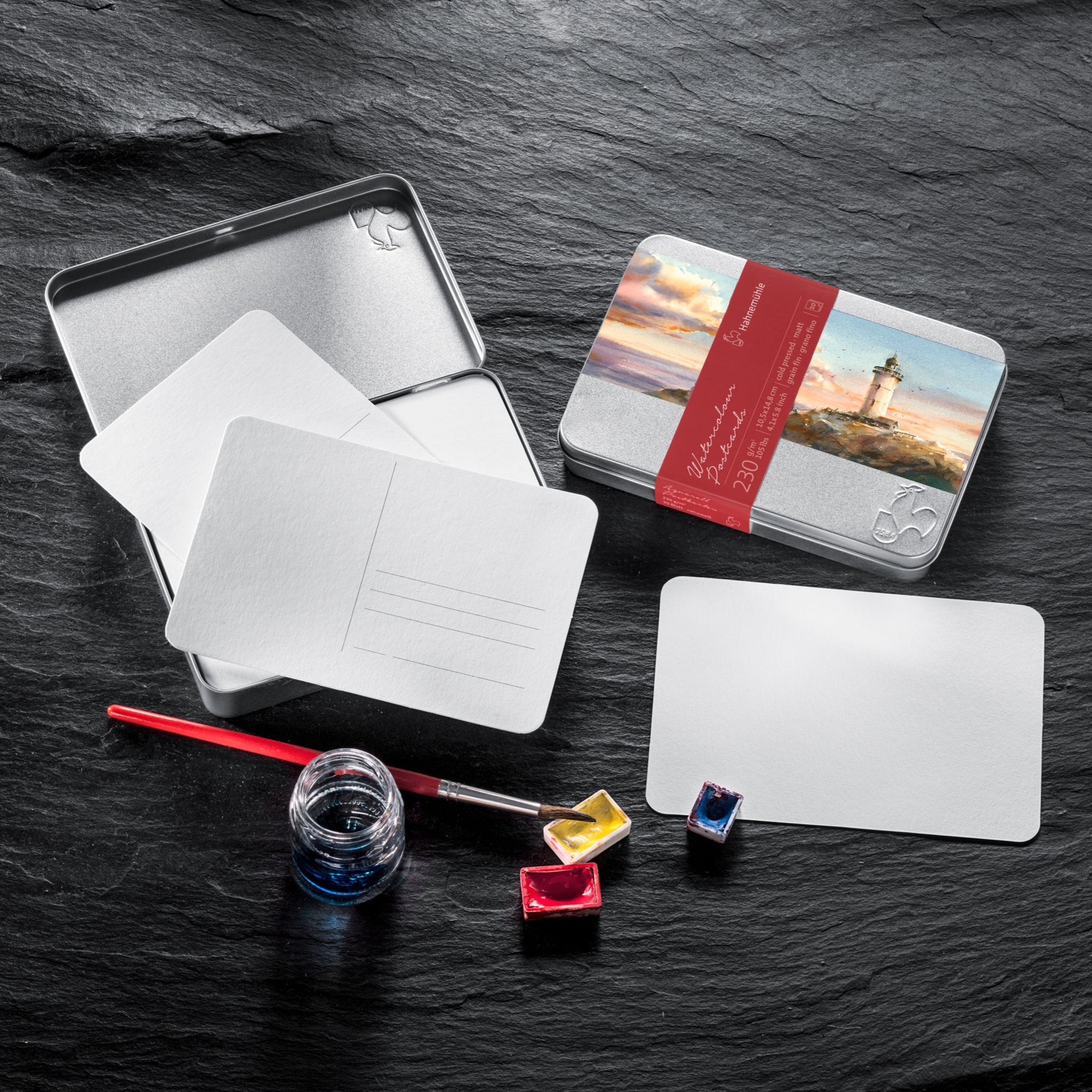 A set of white postcards displayed alongside a brush, ink, watercolour half pans and a metal tin with a red label on a dark slate background.