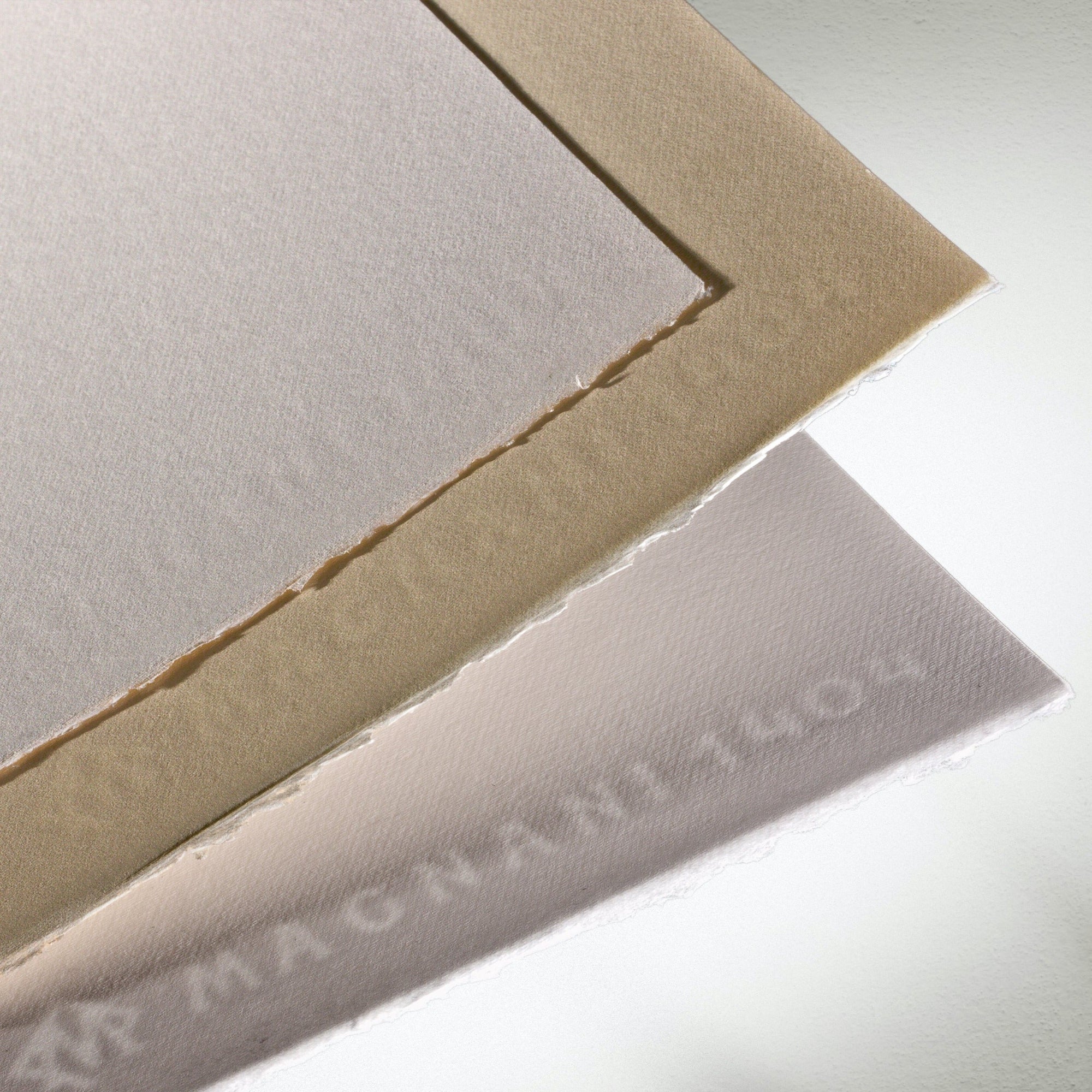 Close up of stacked sheets of lightly textured Magnani 1404 Incisioni paper, with visible watermark, in beige, white and ivory colours.