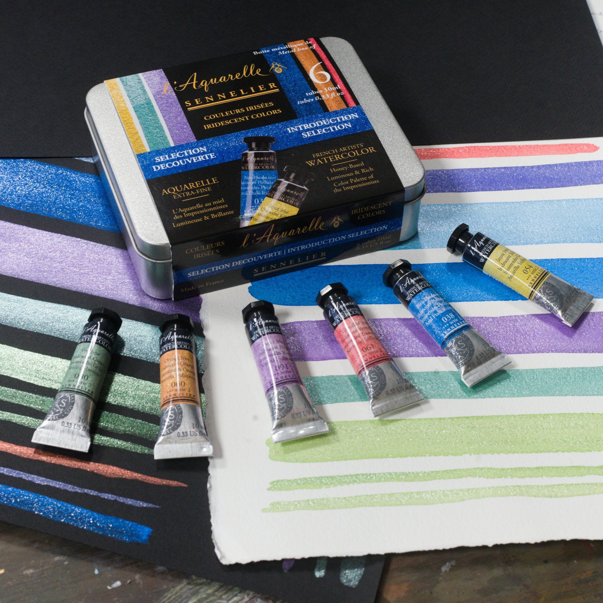 A metal box set of 6 iridescent watercolour paints with the 6 tubes displayed beside it atop test strips on black and white paper of the iridescent watercolours.