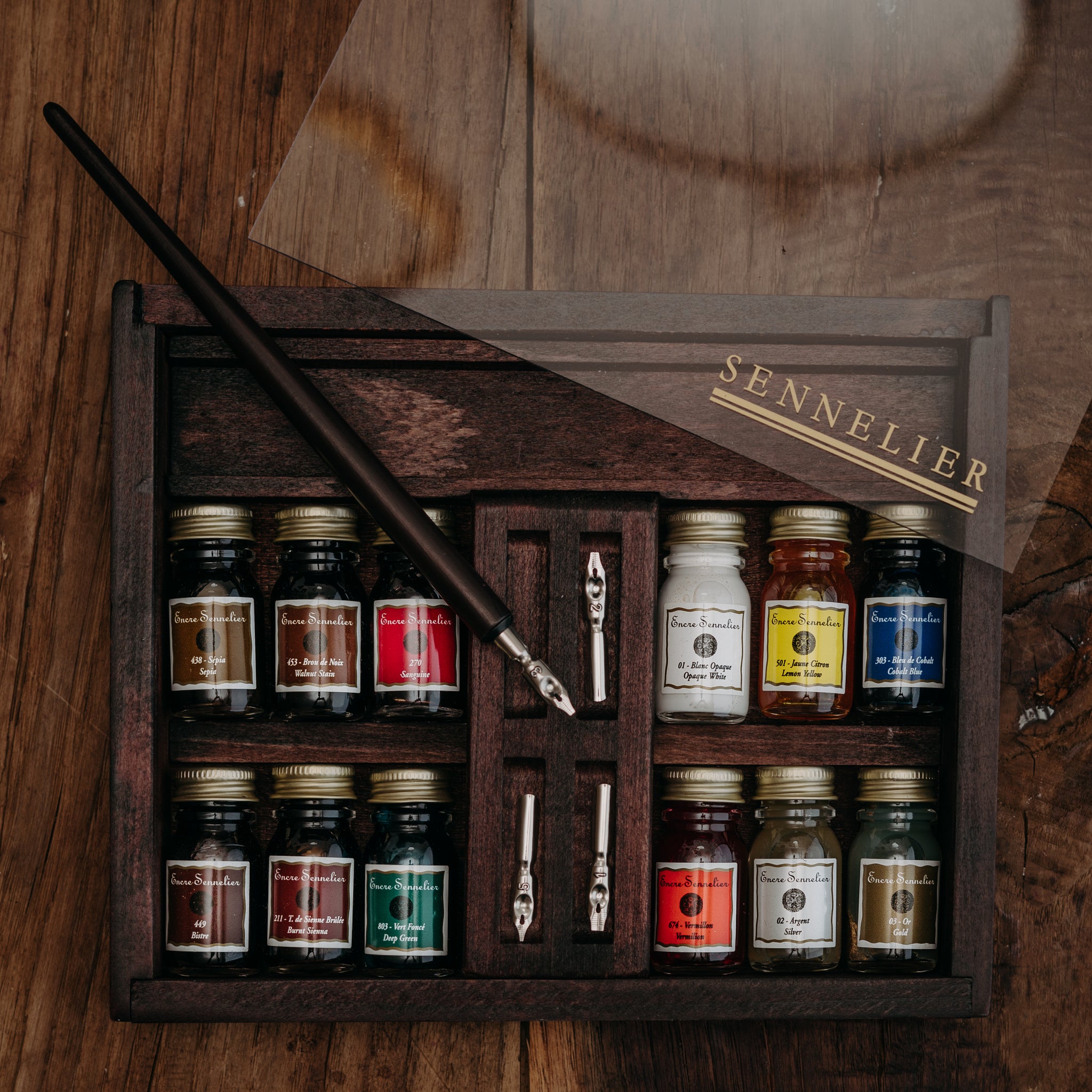 A wooden box set with a clear acrylic lid containing multiple glass bottles of Sennelier ink, several nibs, and a dip pen, sitting on a wooden surface.