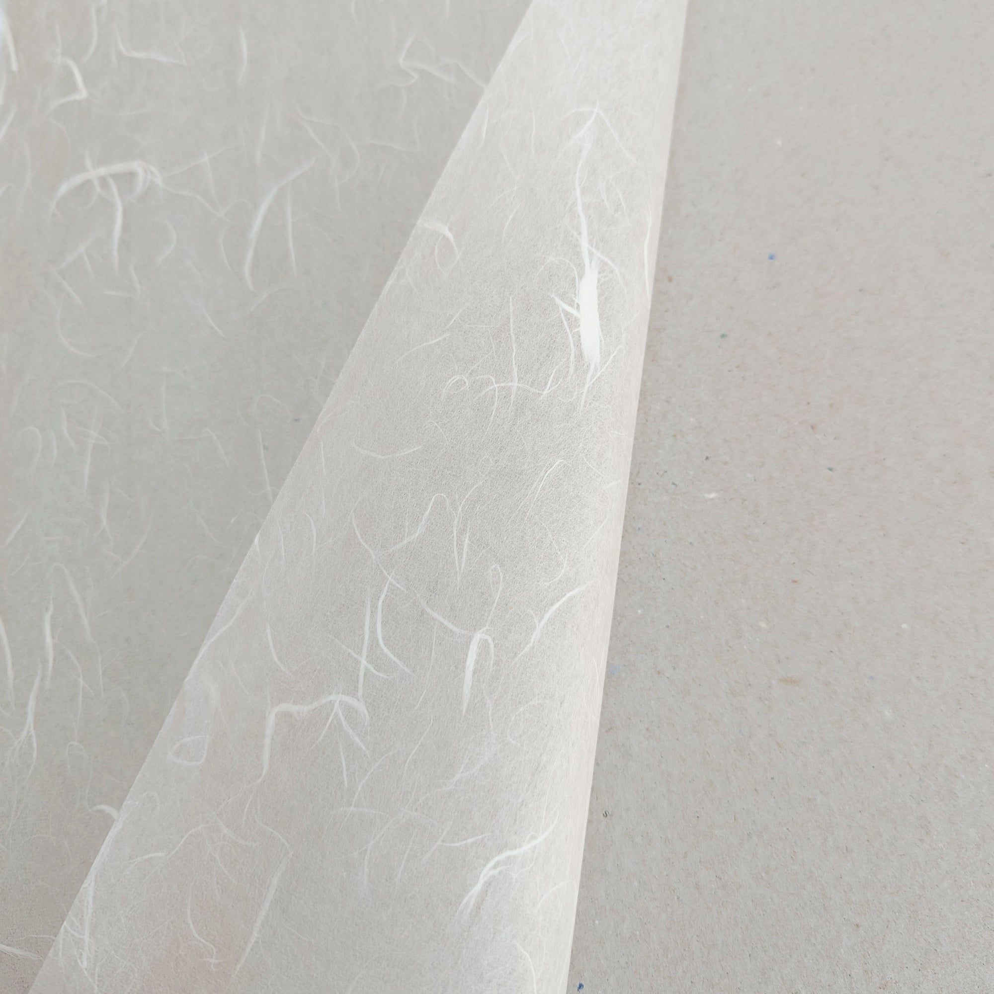 A sheet of Unryu White Light paper with a semi-transparent quality and swirling fibre texture. Shown turned over on itself to show both sides and placed against a grey background.