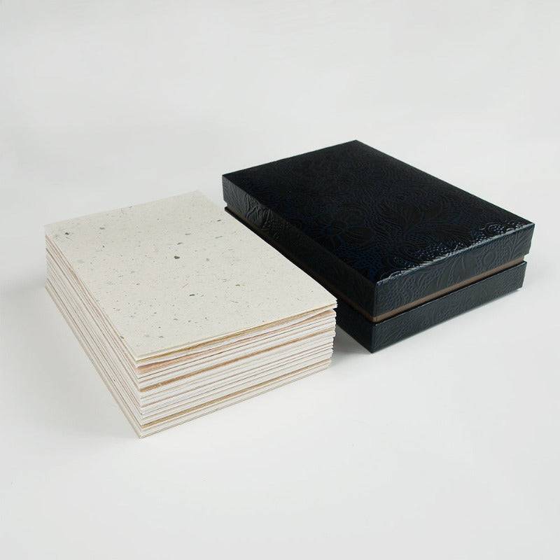 A black lacquered box with a floral pattern shown next to a pile of various Japanese papers in neutral tones