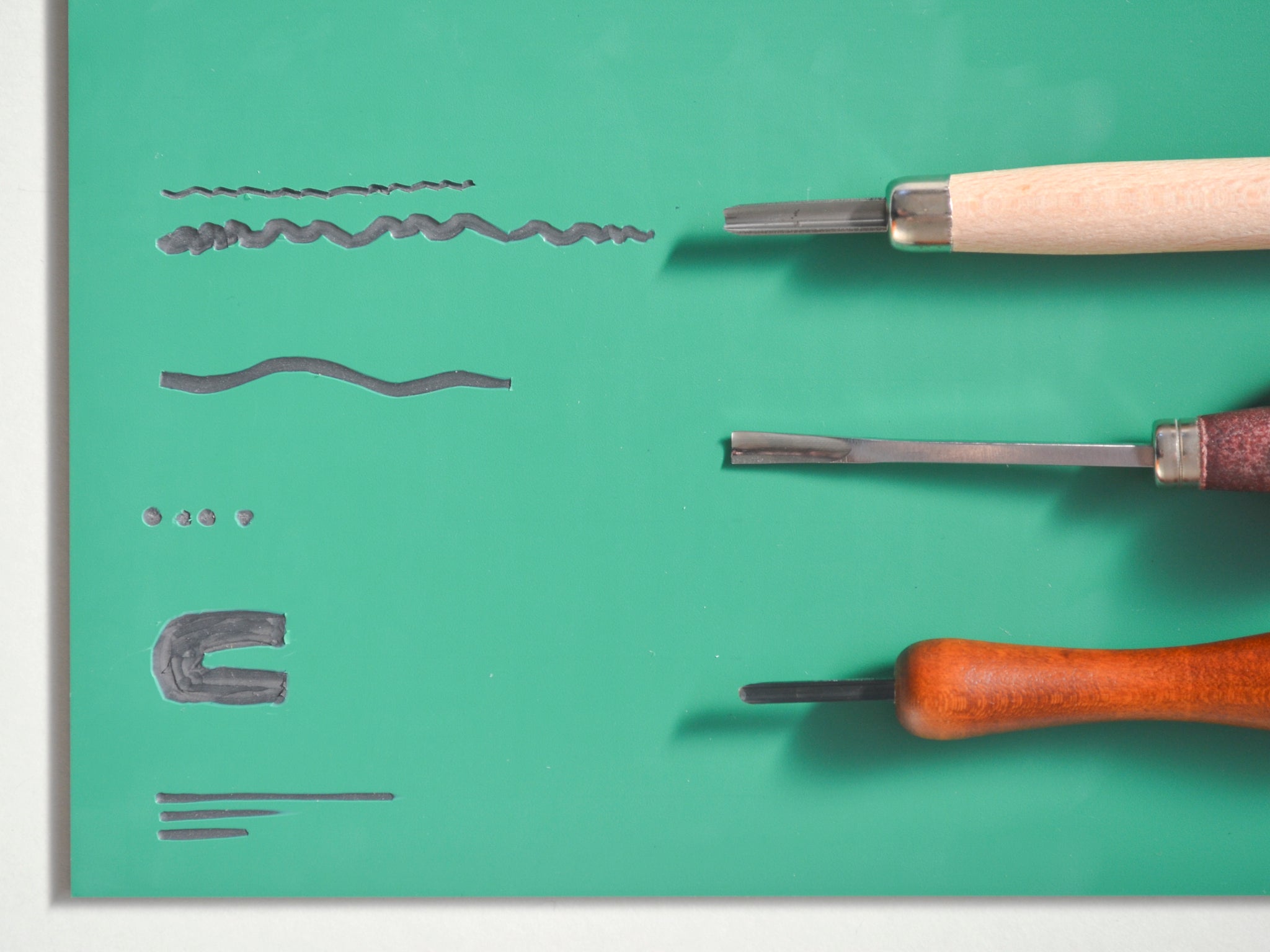 Trio of U cutting lino tools on a green vinyl tile with lines and patterns made by the tools.