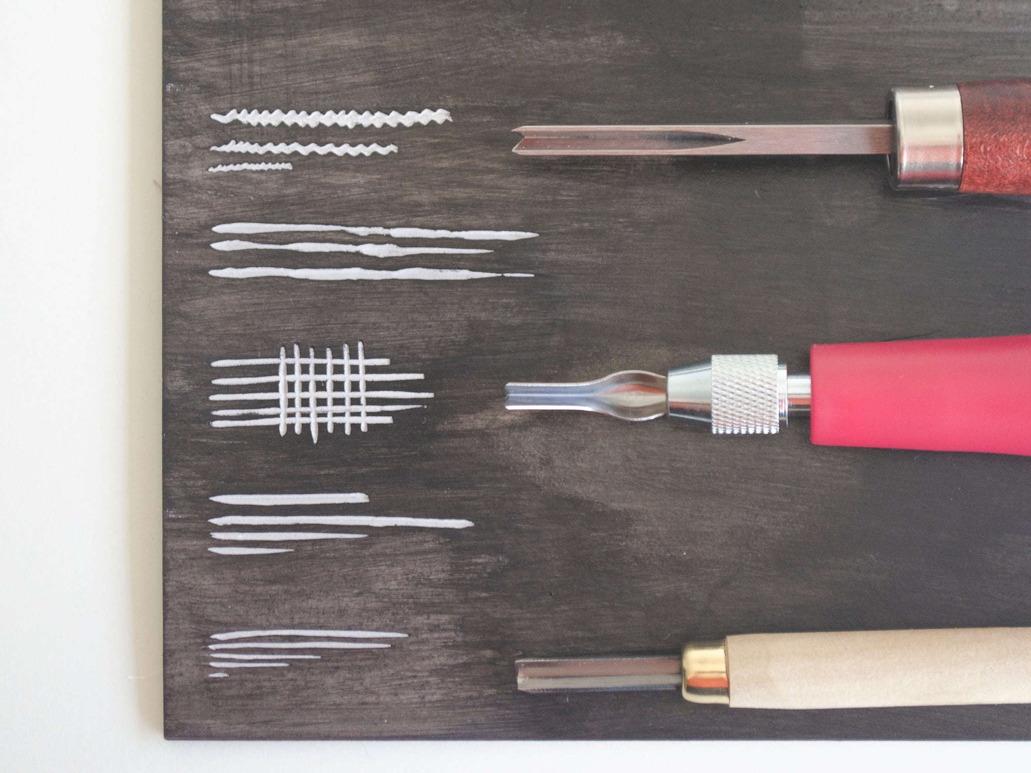 trio of V cutting lino tools on a stained piece of lino with lines and patterns made by the tools.