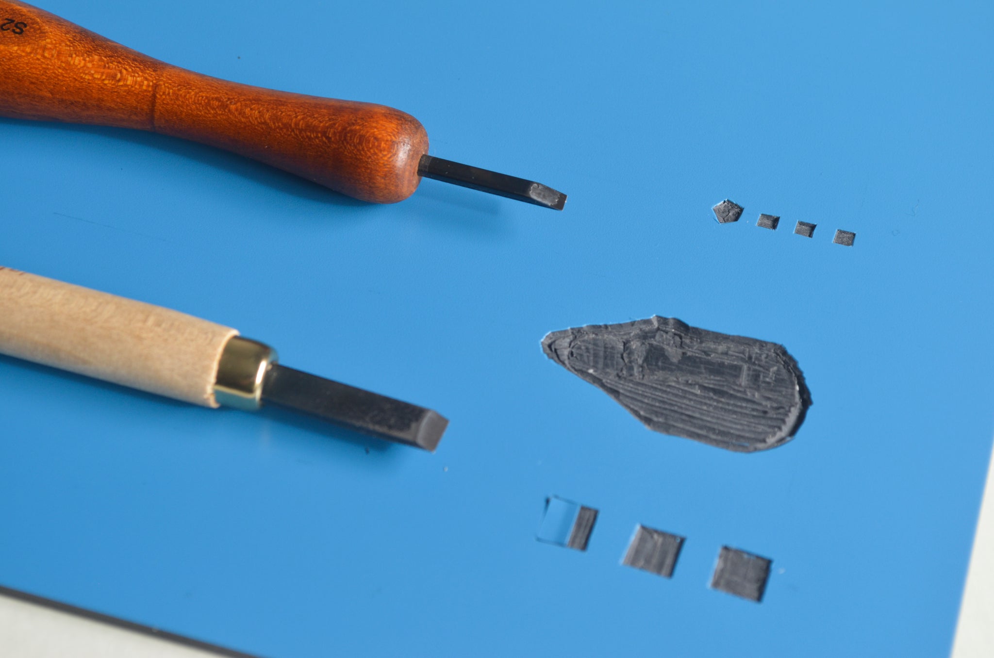 Chisel cutting lino tools on a stained piece of lino with lines and patterns made by the tools.