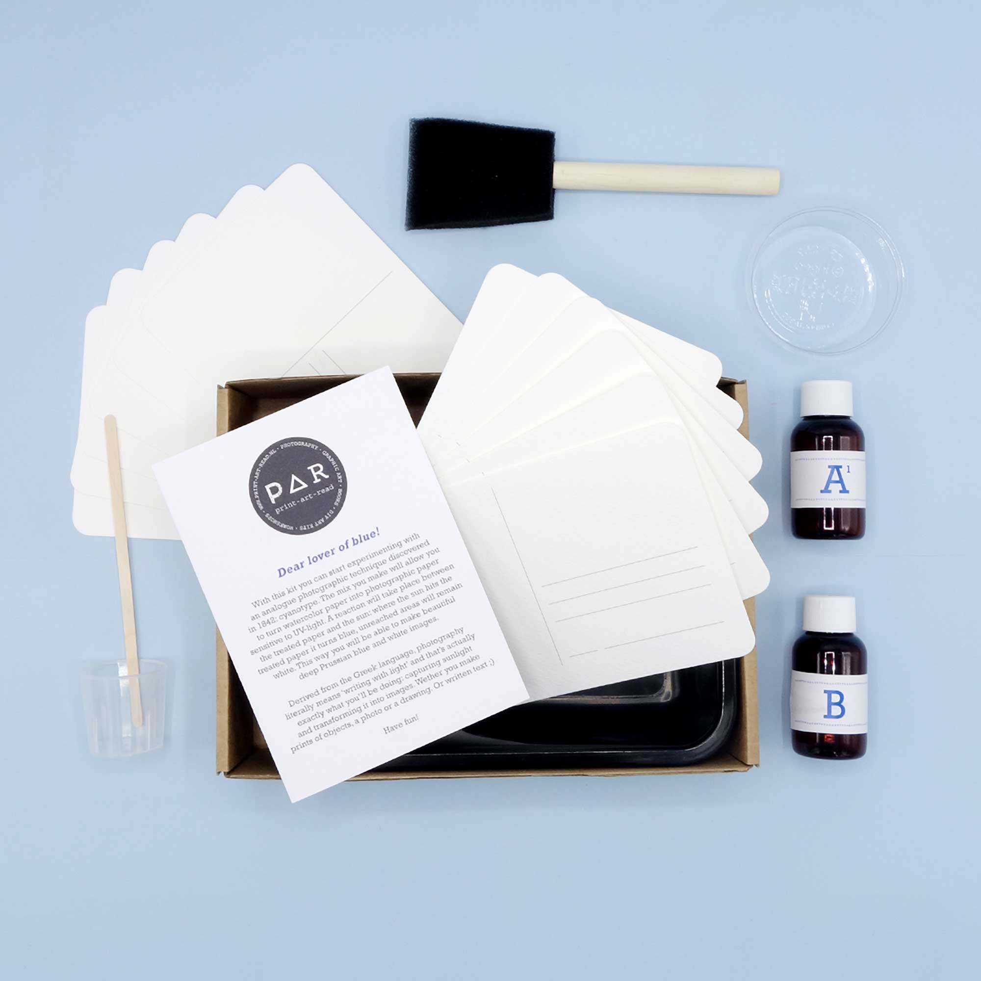 Cyanotype Postcard Kit contents including; postcards, 2 bottles labelled A and B, a Foam Brush, trays and some plastic cups. Displayed on a blue background