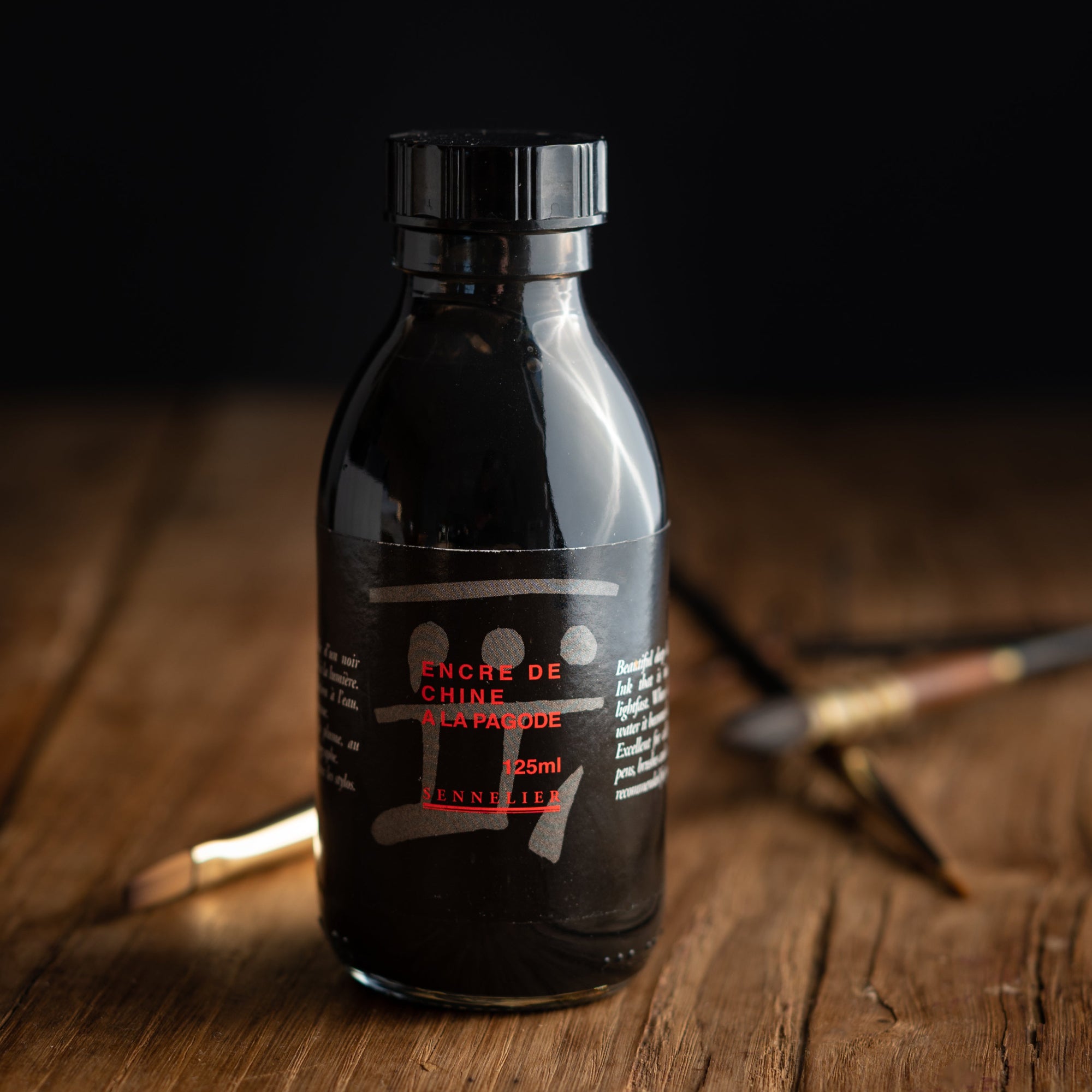 A 125ml Bottle of Sennelier Pagode Black Encre Indian Ink on a wooden table with brushes sitting in the background.