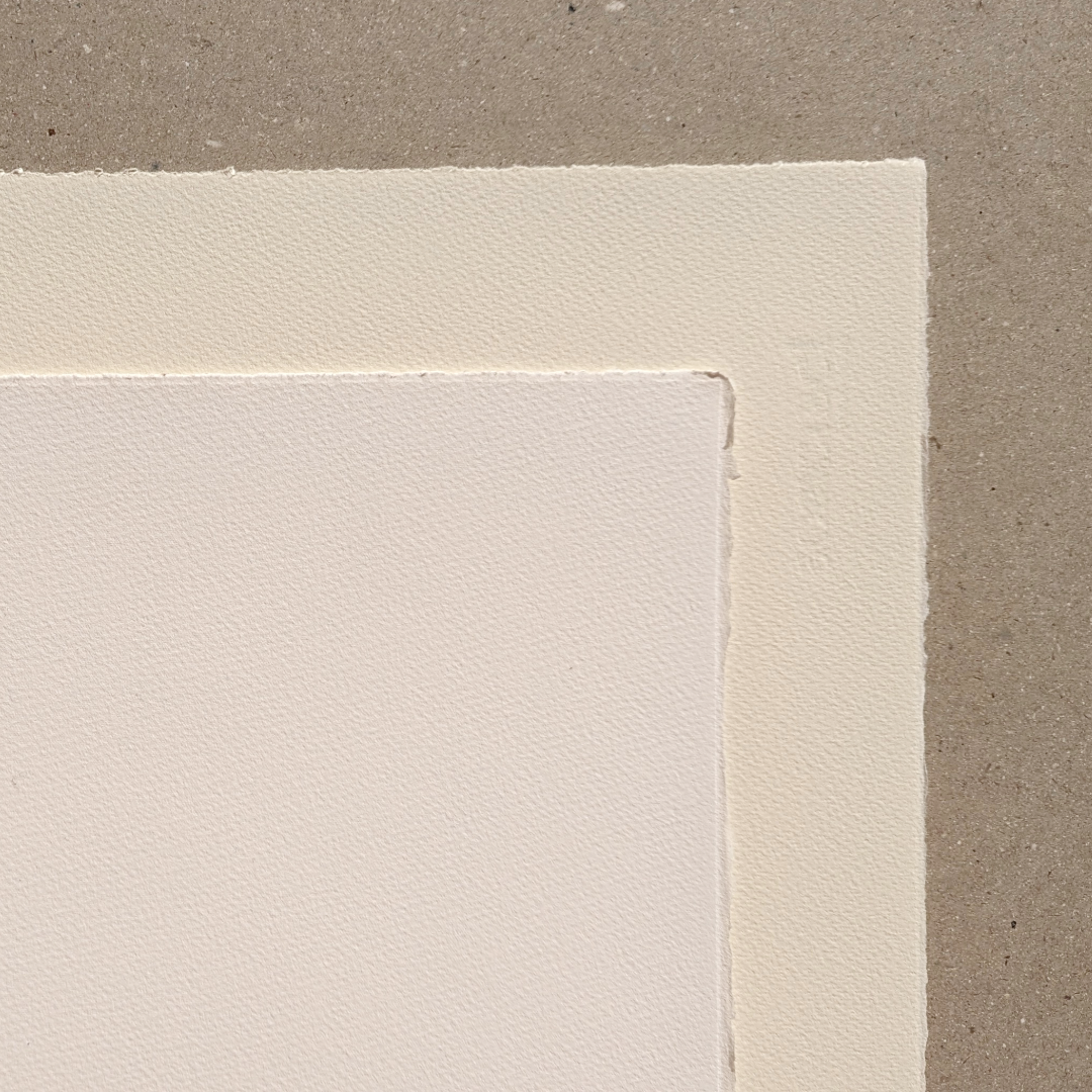 A close up of two sheets of textured Somerset papers, one in white and the other in an off-white tone. Shows the decide edges of the paper on a grey-brown background.