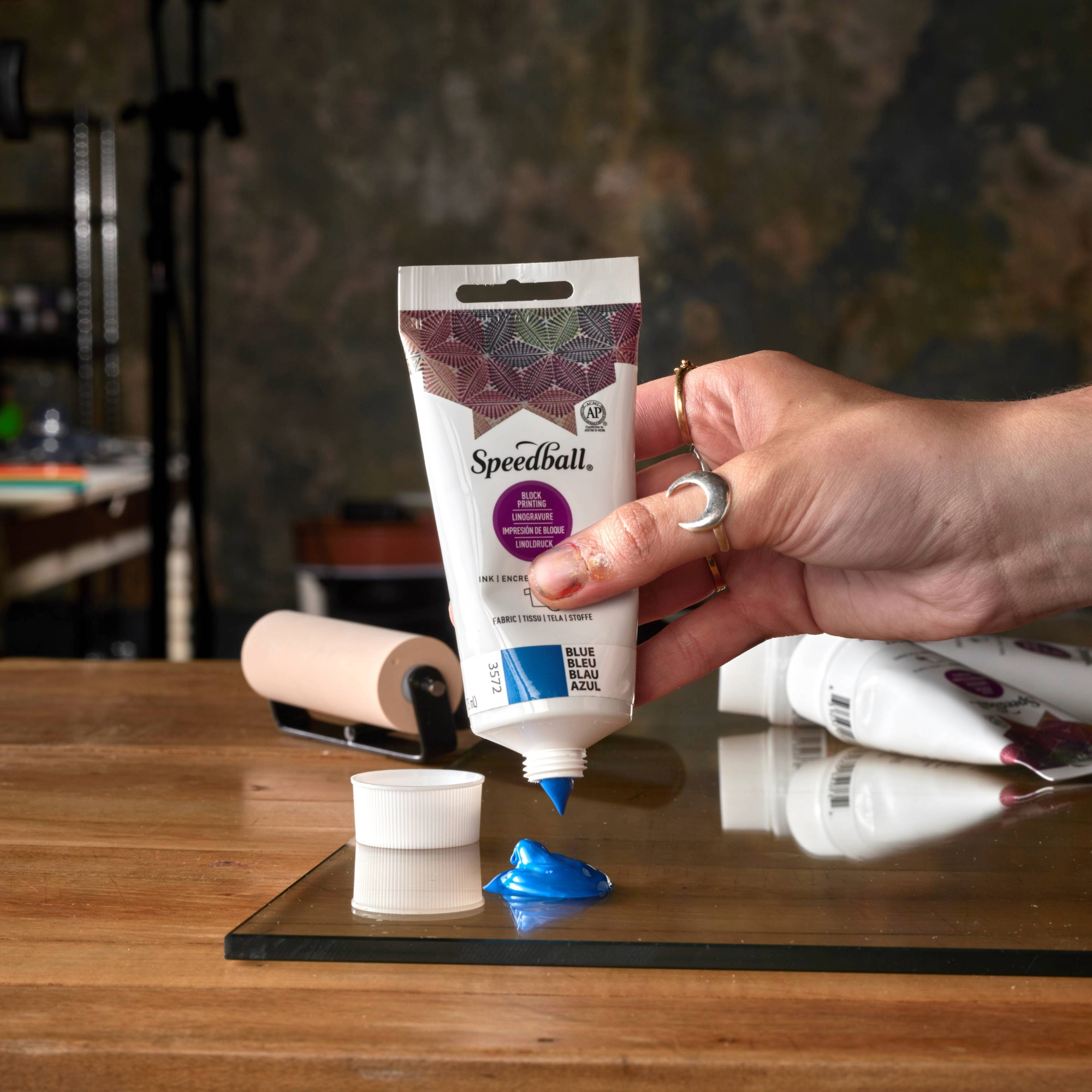 Hand holding a Speedball Fabric Block Printing tube with blue ink on a glass slab with a roller and wooden table in the background.