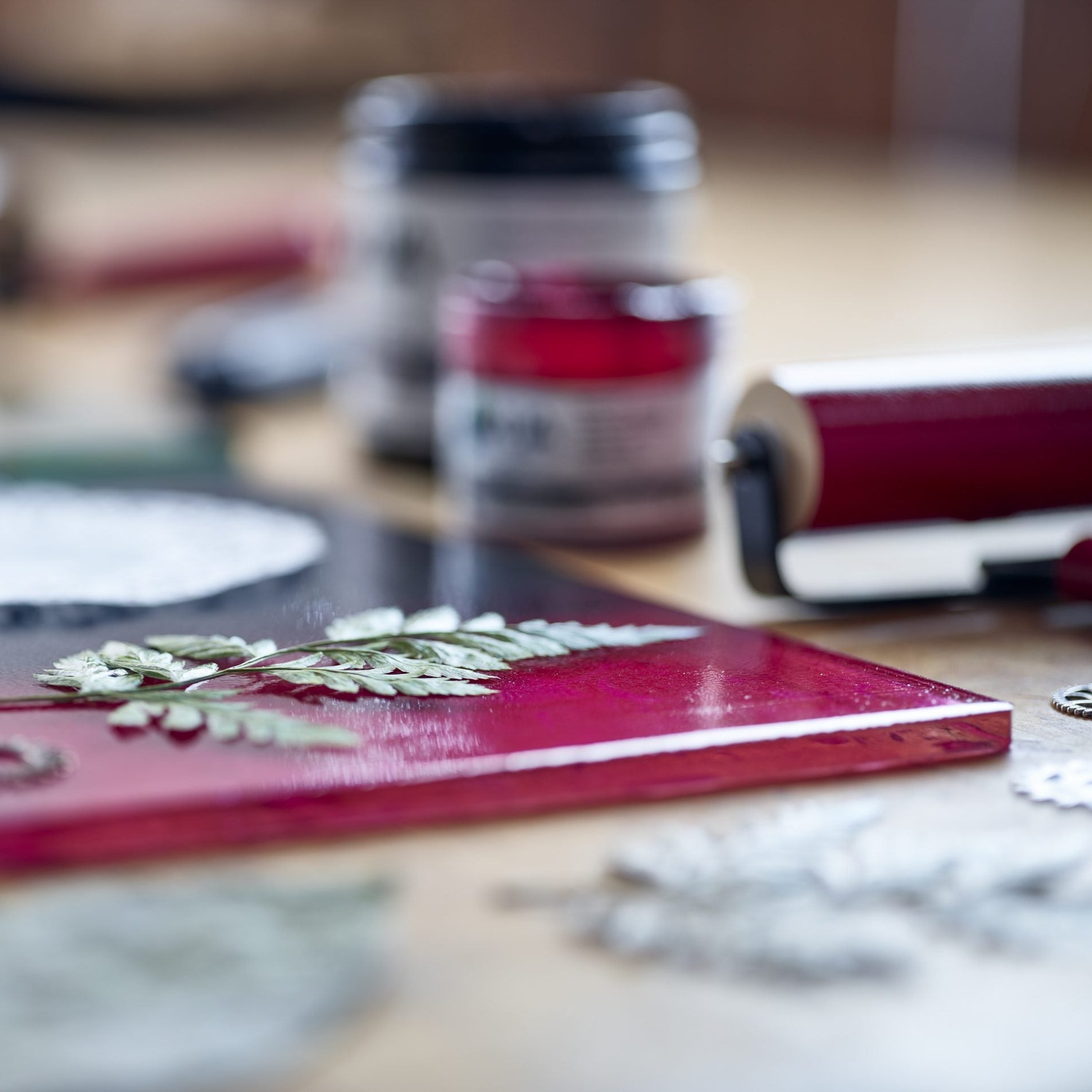 Gel printing plate inked up with red ink with a leaf sitting on top next to a inked up roller with Akua ink tubs in the background.