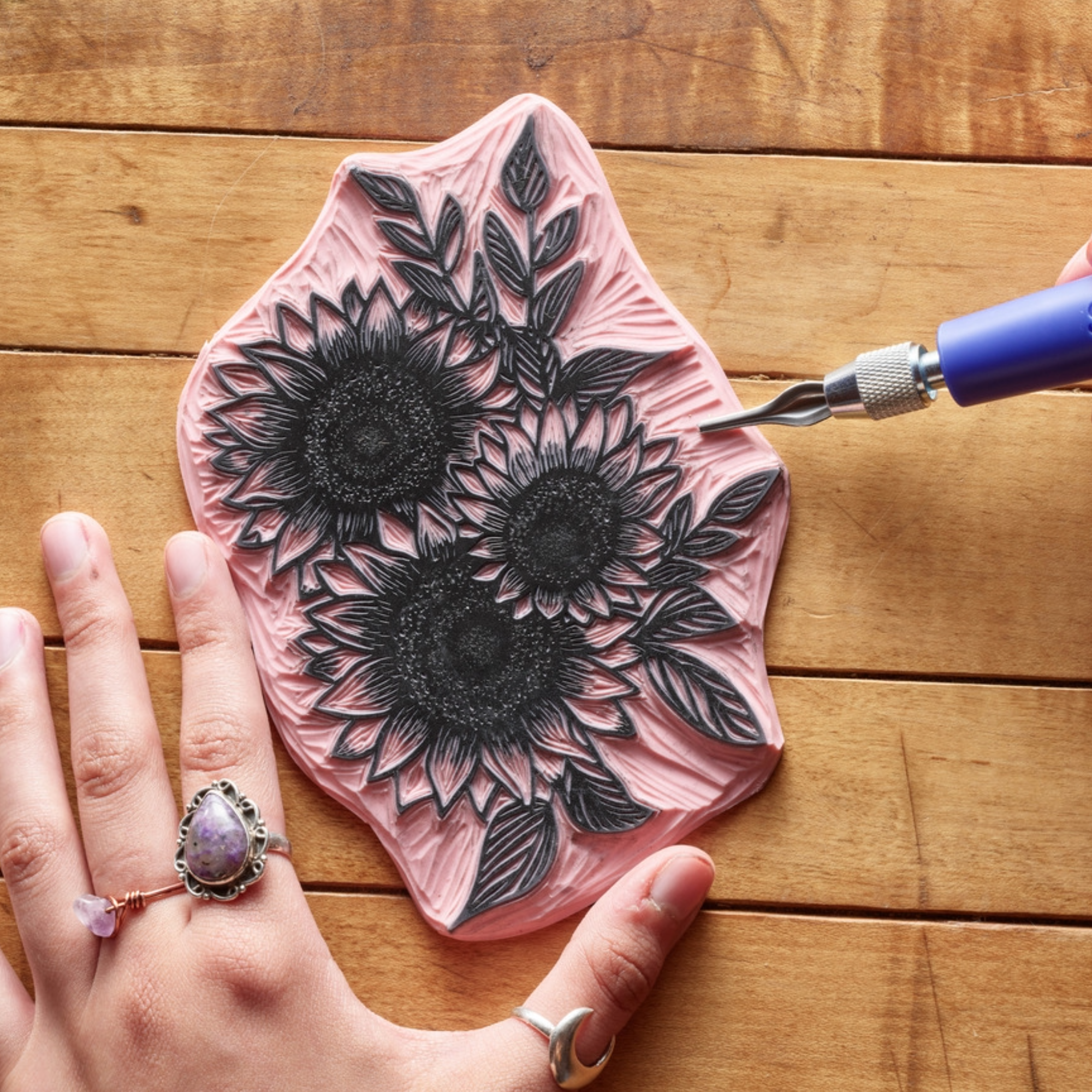 Hand holding a pink speedy carve block with a black floral design, being carved using a speedball tool on a wooden surface.