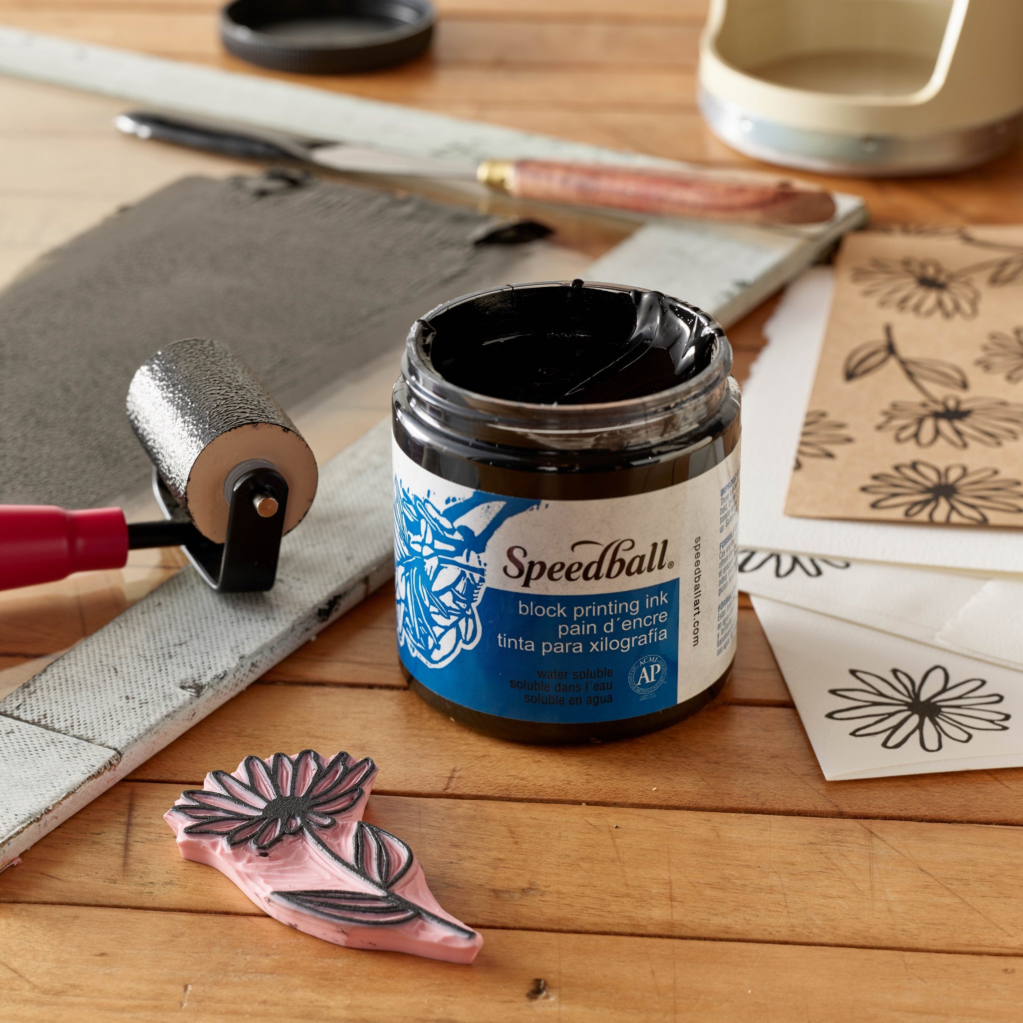A jar of Speedball Water Based Block Printing Ink in black, with a label showing the brand and product name, placed on a worktable with a variety of block printing tools and materials, including a roller, papers with printed designs, and a carved block.