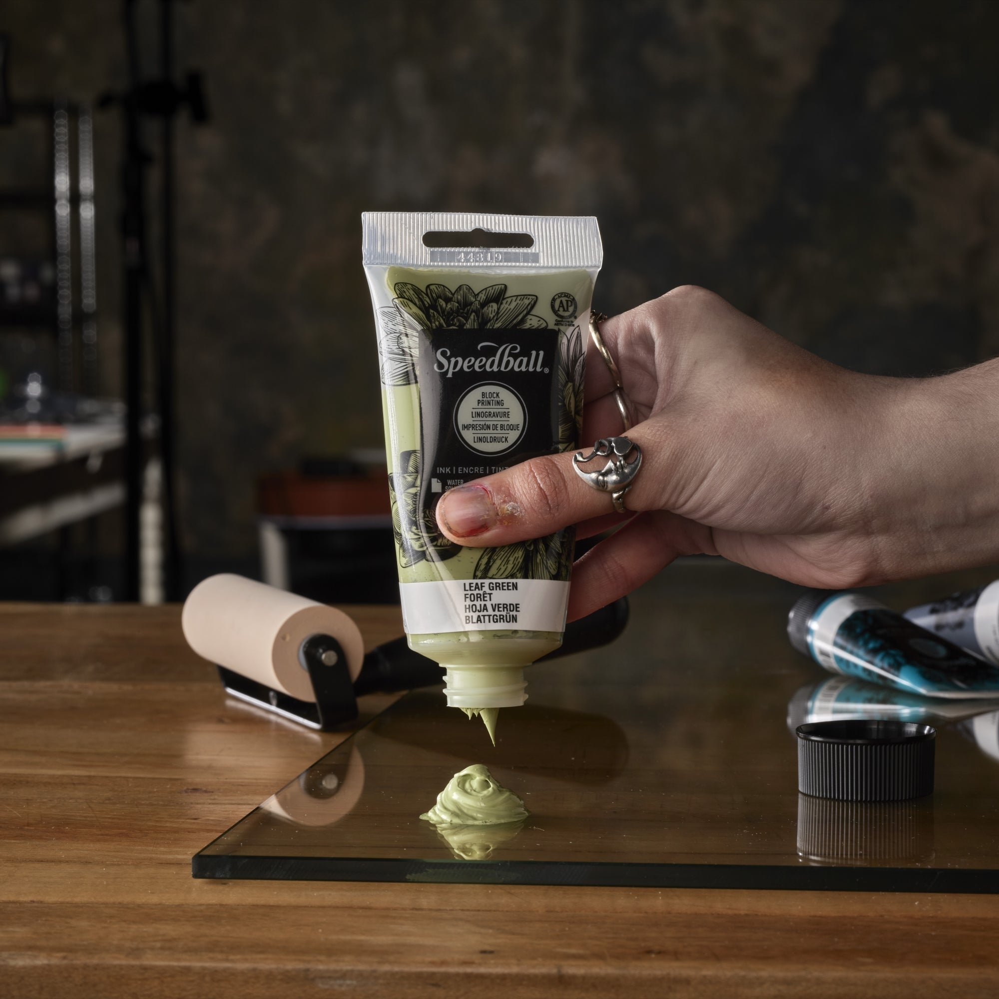 Hand squeezing a Speedball paint tube over a glass slab on a wooden table with a blurred background