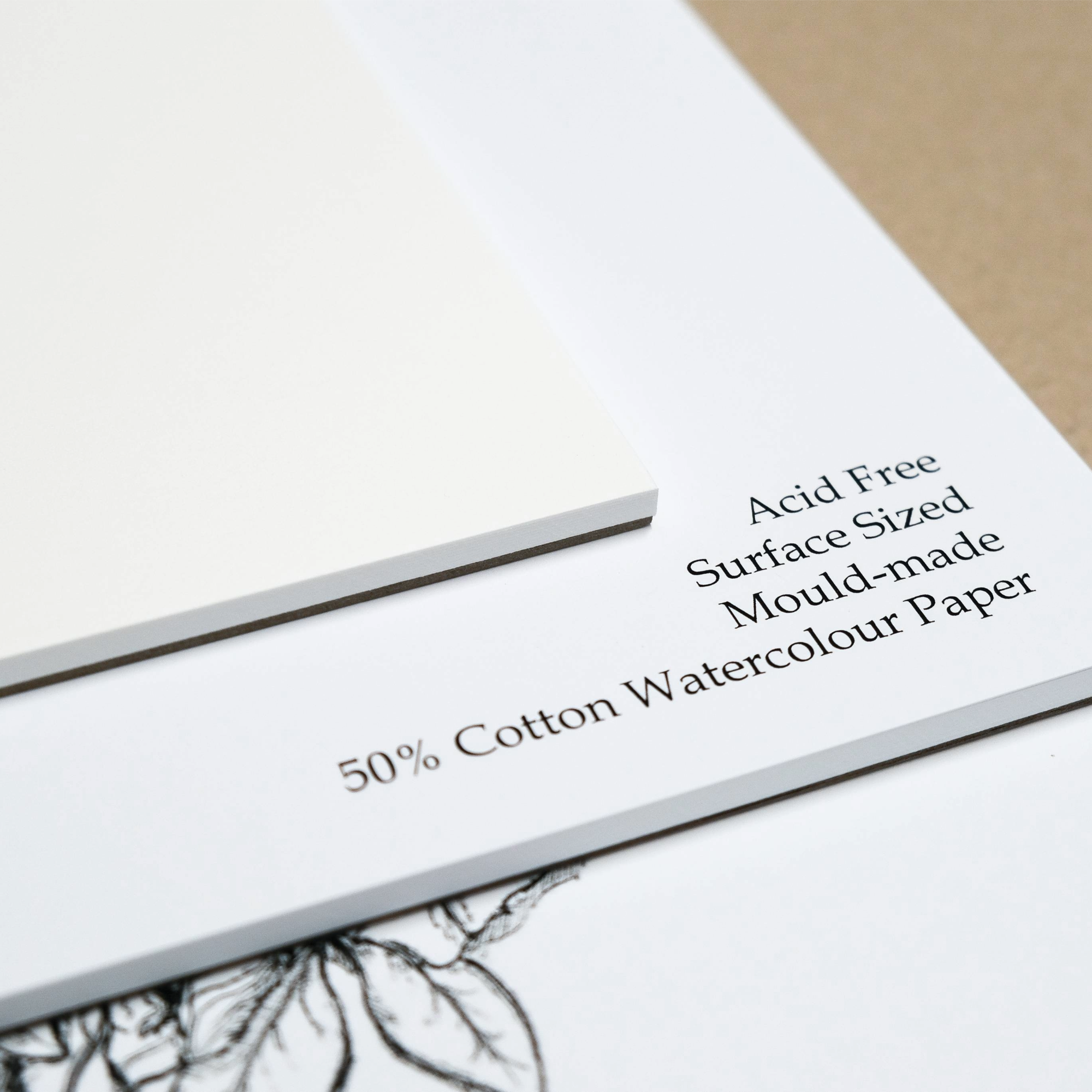 Close up of a stack of paper pads with the top pad showing the surface of the smooth white paper. The second pad list the features of the paper: 'Acid Free, Surface Sized, Mould-made and 50% Cotton Watercolour Paper.