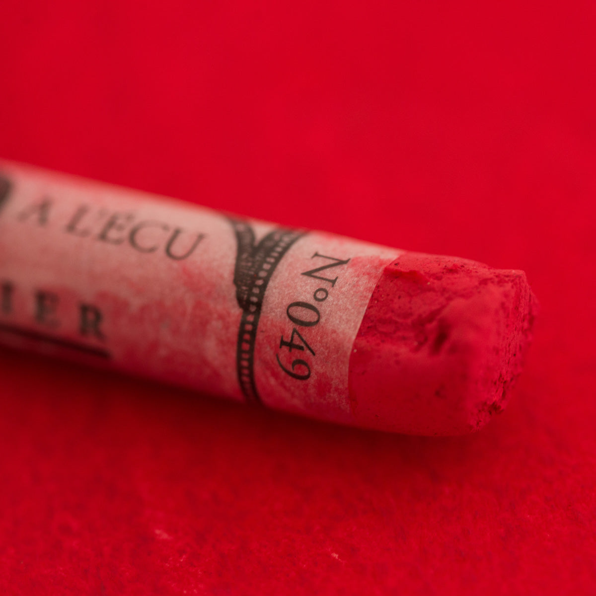 A close-up of a red Sennelier extra soft pastel on the same coloured background, with the colour information printed on the paper wrap.