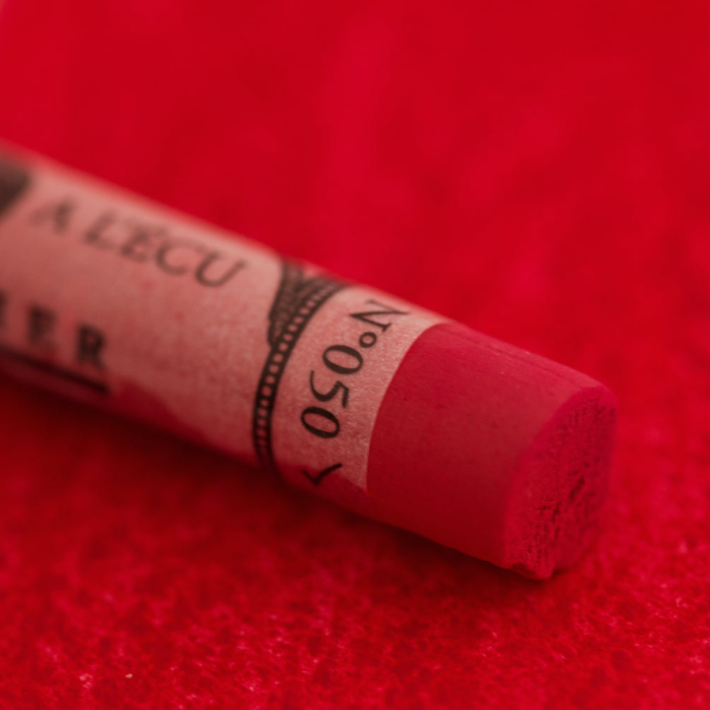 A close-up of a red Sennelier extra soft pastel on the same coloured background, with the colour information printed on the paper wrap.
