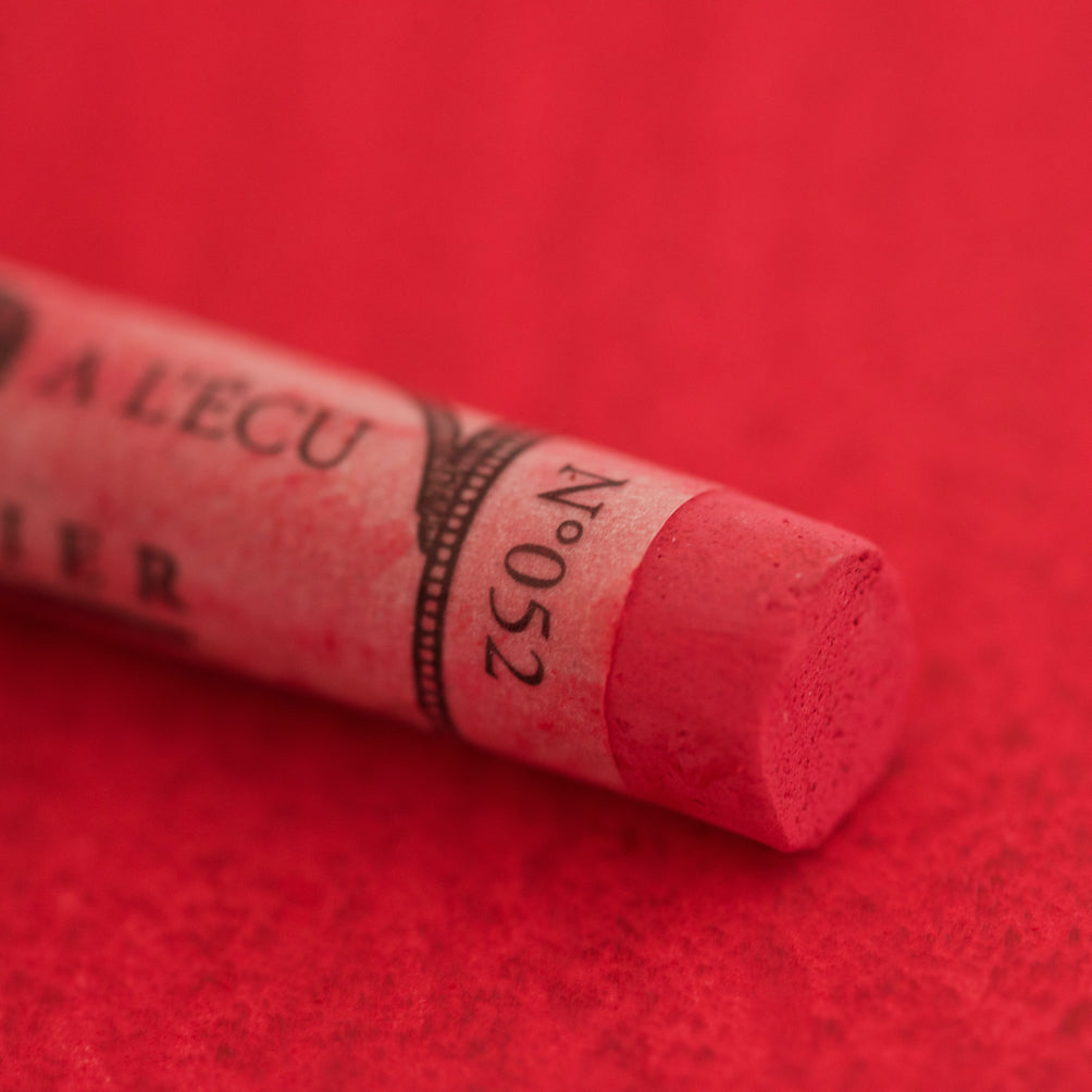 A close-up of a red Sennelier extra soft pastel on the same coloured background, with the colour information printed on the paper wrap.