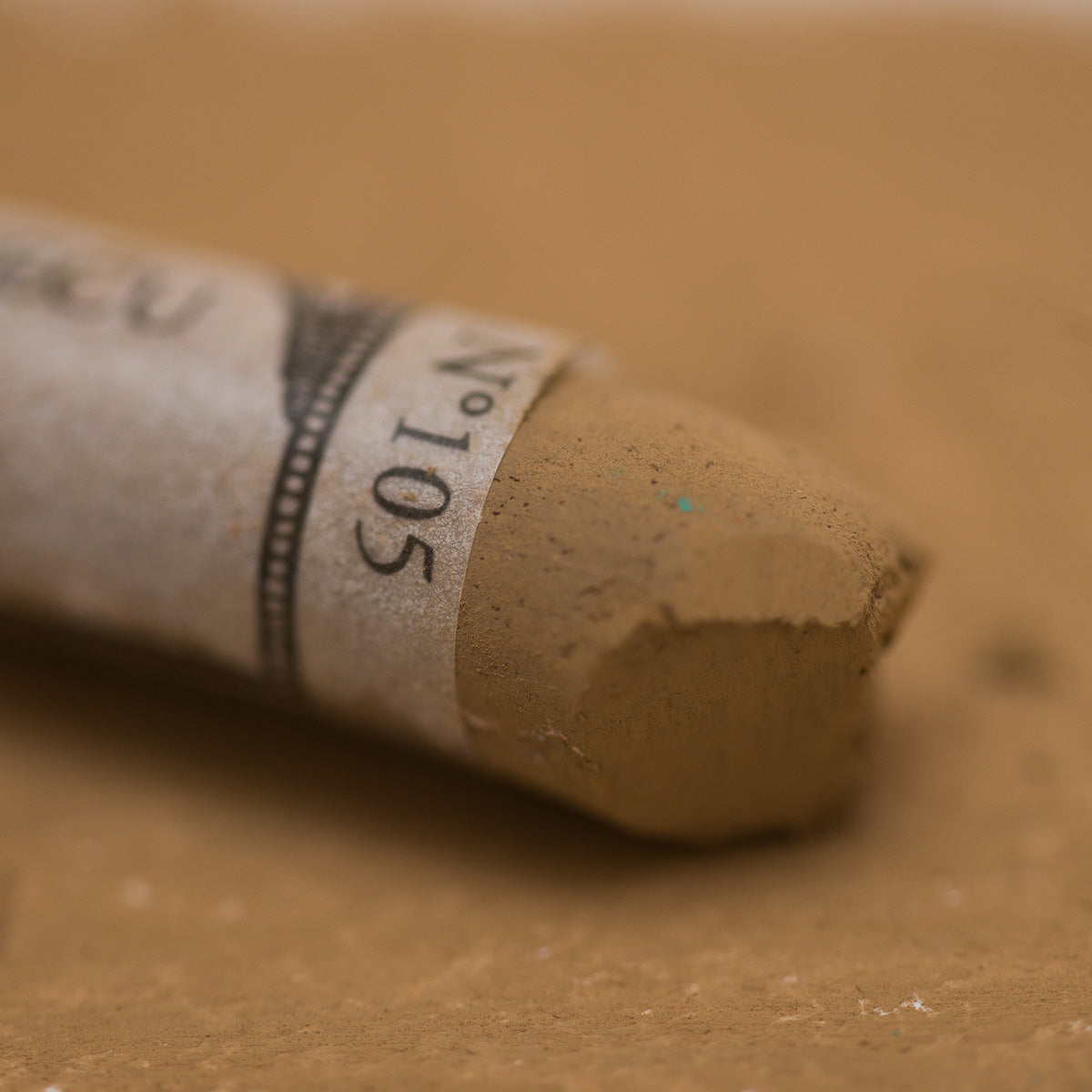 A close-up of a brown Sennelier extra soft pastel. on the same coloured background, with the colour information printed on the paper wrap.